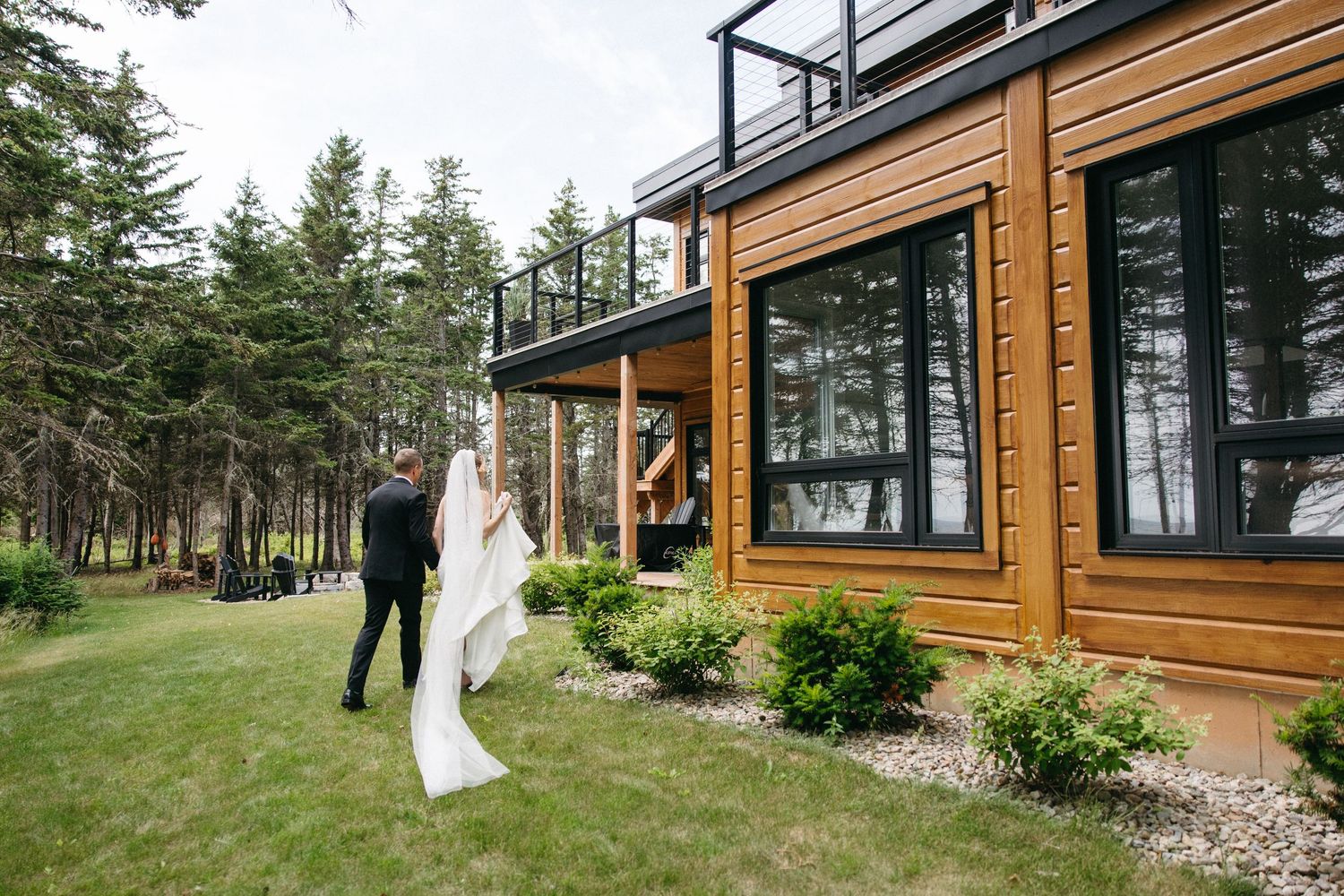 Bride in flowing white gown and veil walks with groom beside modern wood-clad venue.