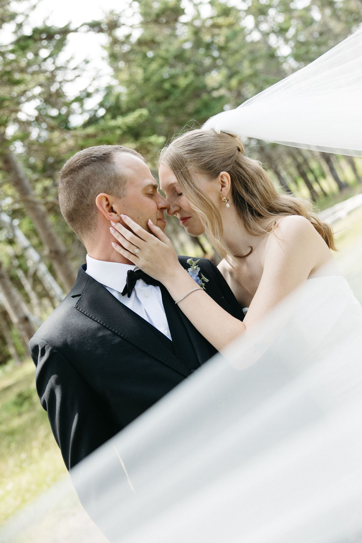 Bride tenderly touches grooms face as they share romantic moment under flowing white veil outdoors.