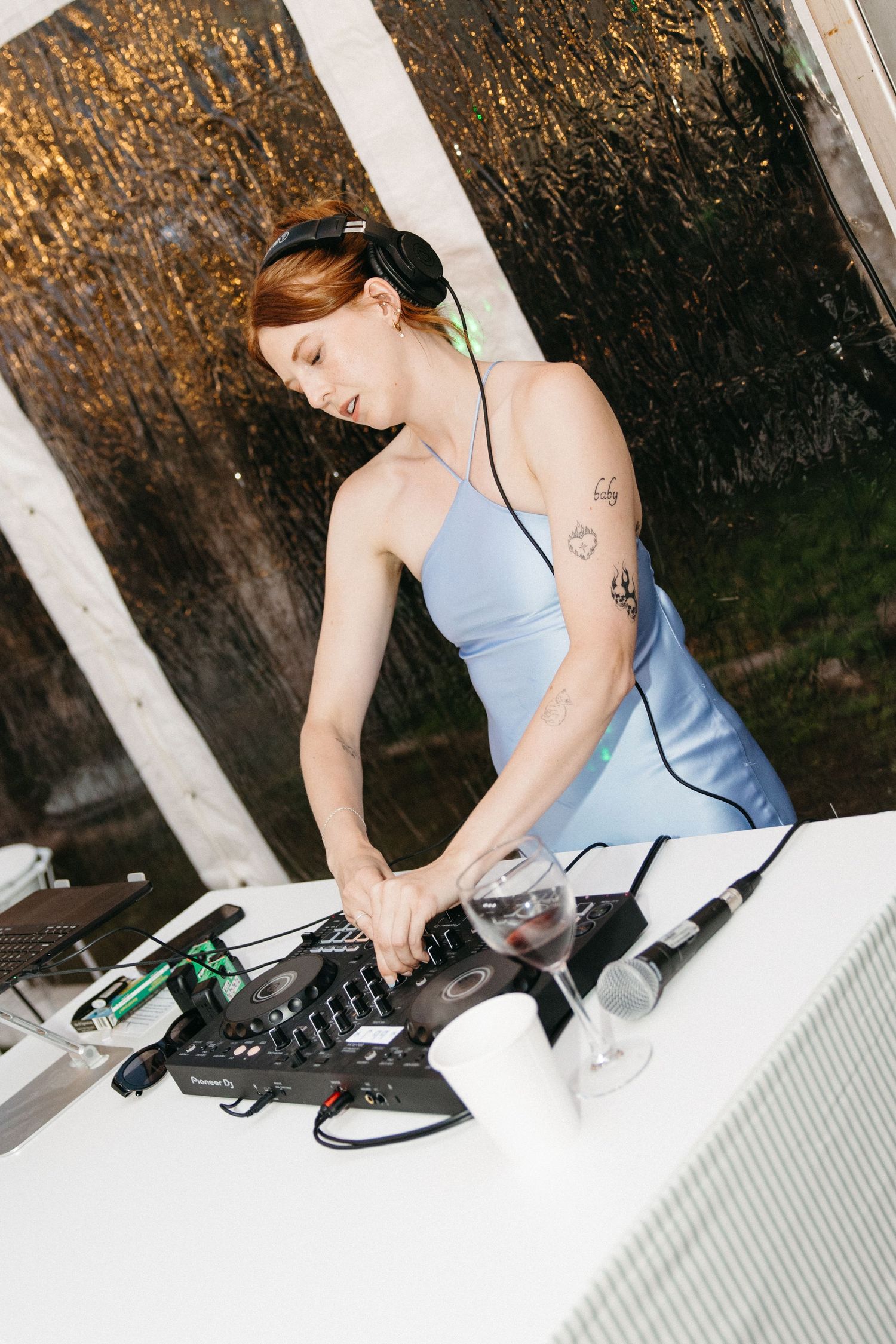 Female DJ with tattoos and headphones mixing music at outdoor wedding reception in blue dress.