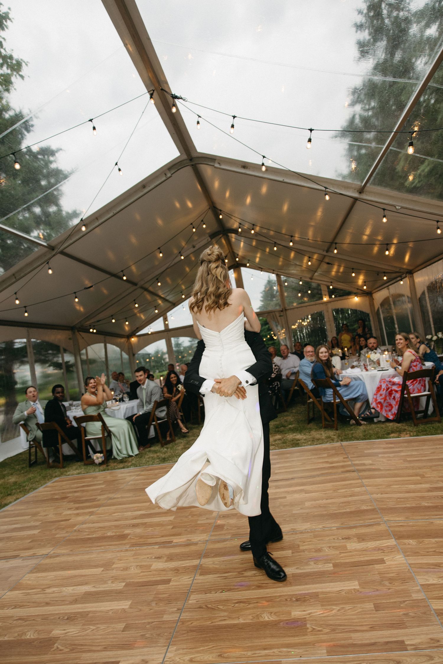 Bride and groom share first dance under string lights in clear wedding tent.