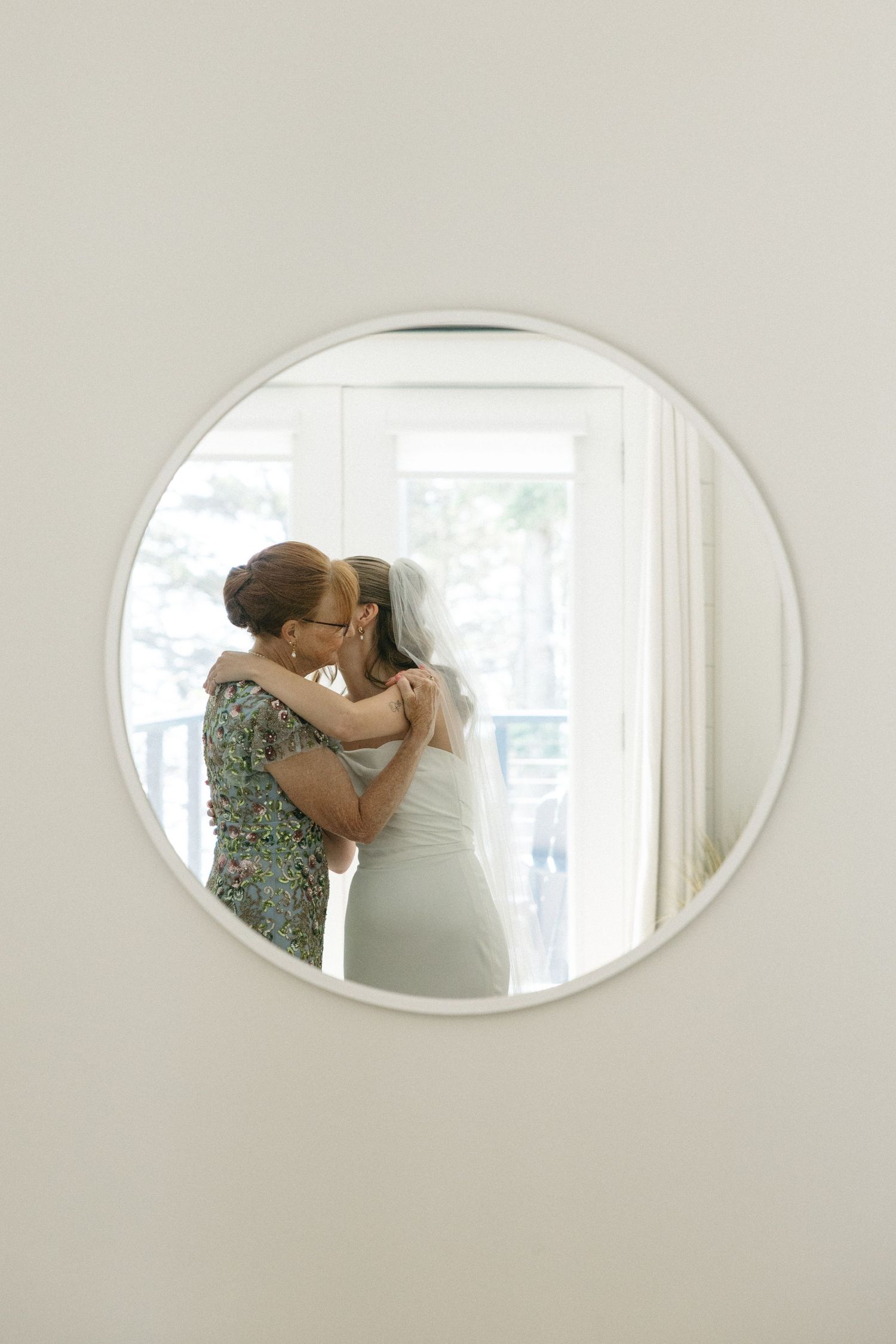 Mother and bride share tender embrace reflected in round white-framed mirror before wedding.