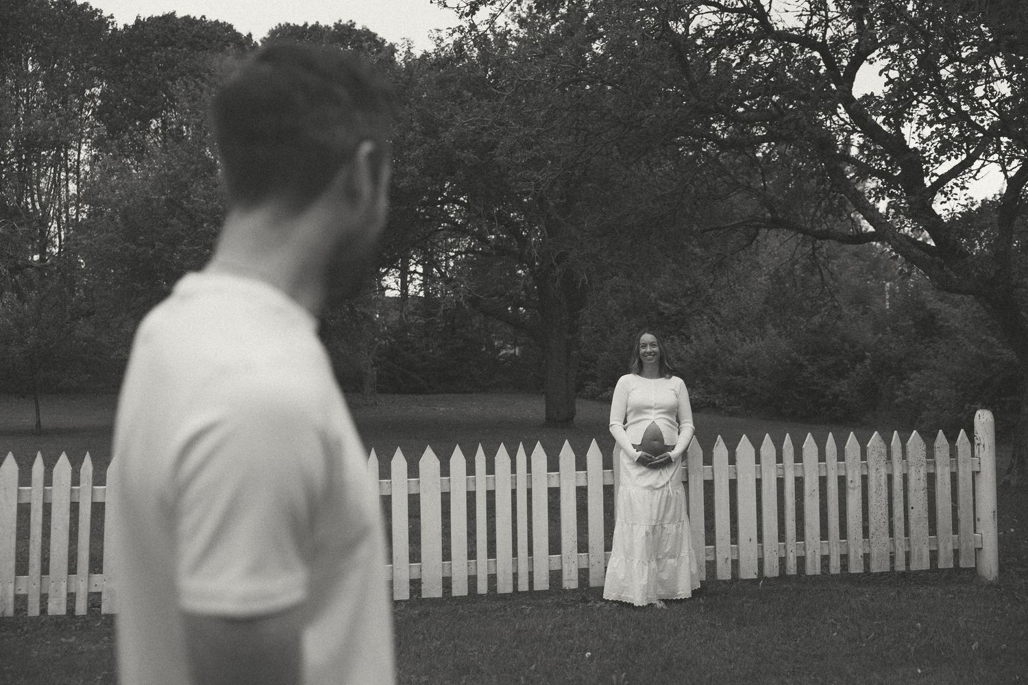 Black and white photo of pregnant woman standing by white picket fence as man looks on in lush garden.