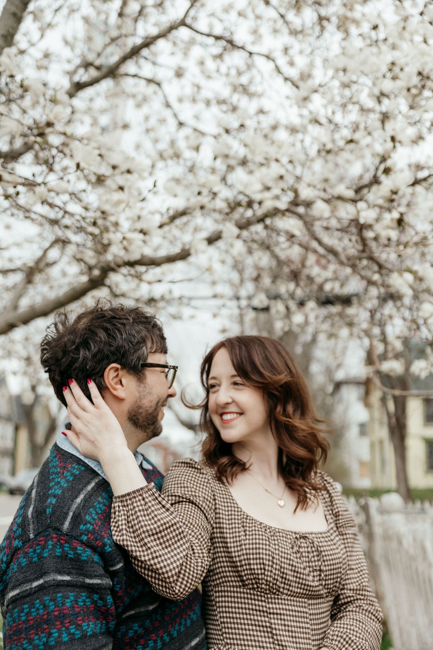 A smiling couple sharing a tender moment under flowering white magnolia trees, the woman touching the mans face.