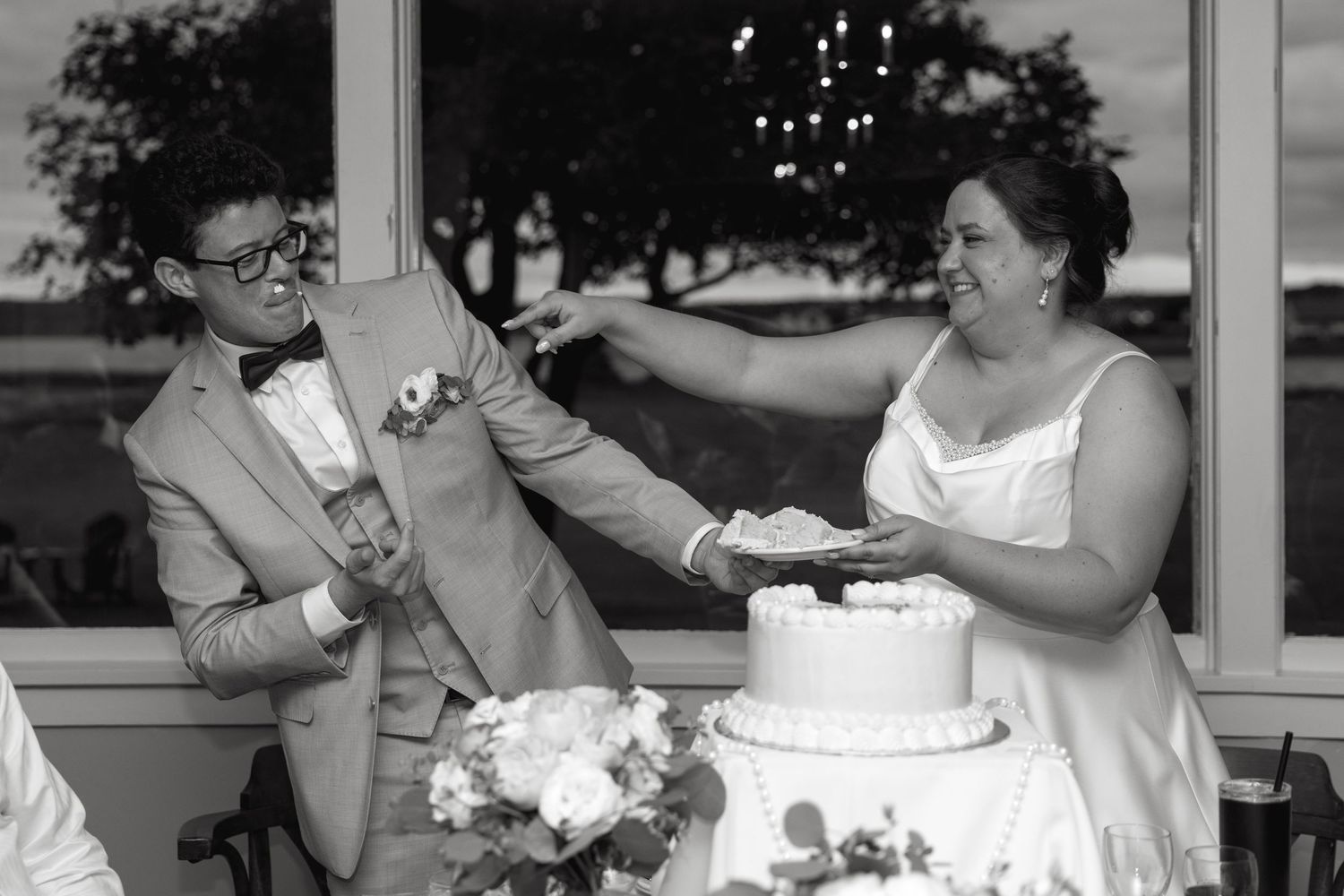 Black and white photo of bride and groom sharing a playful wedding cake cutting moment.