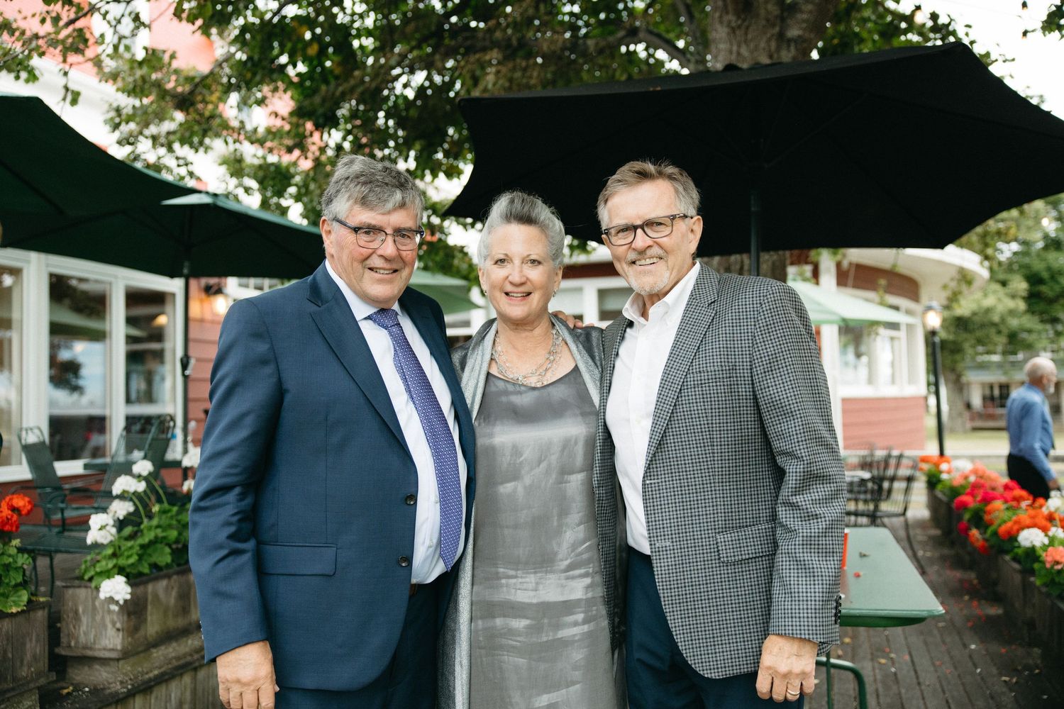 Two men in suits and a woman in silver dress pose together at an outdoor wedding event.