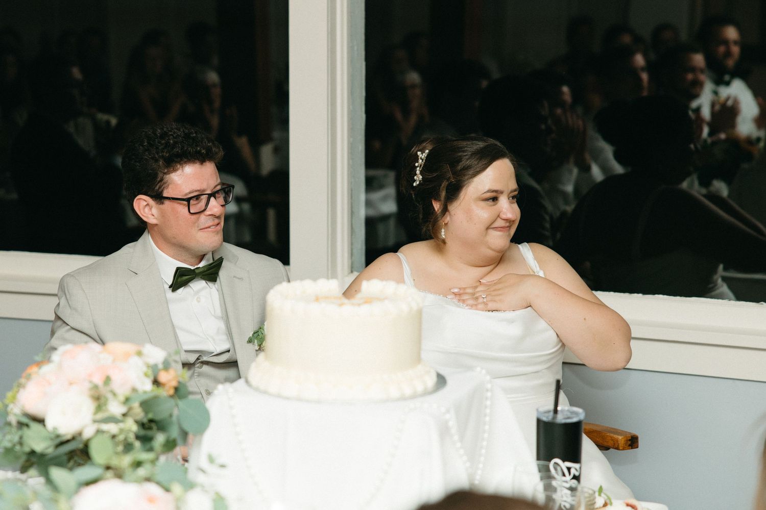 Bride and groom in gray suit sit together at reception table with white wedding cake in foreground.