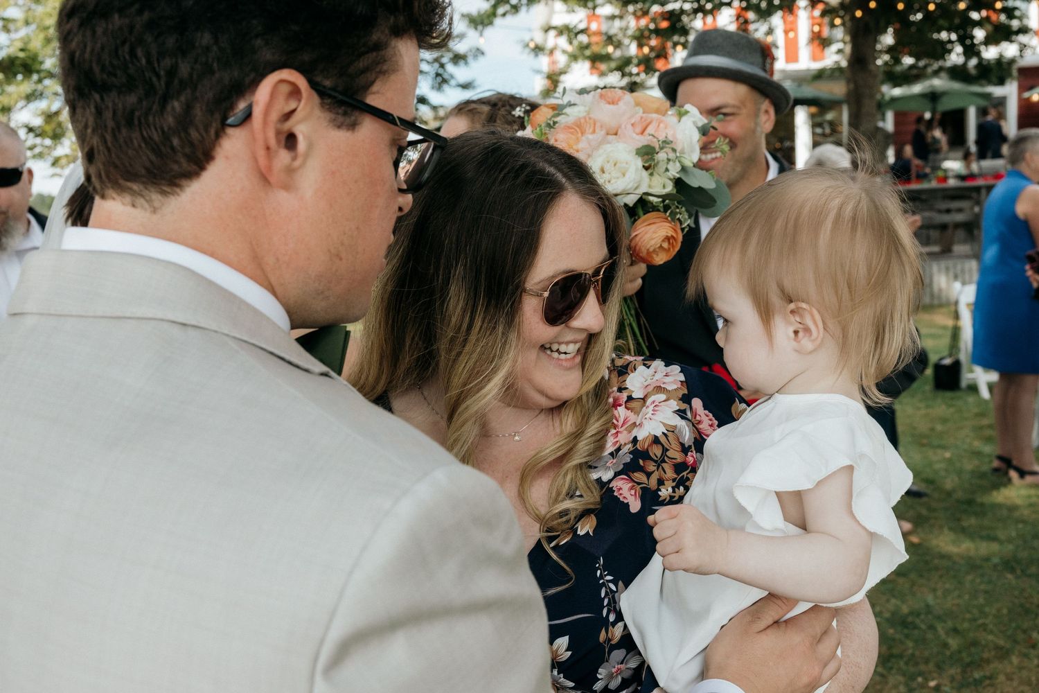 Woman in floral dress holding a baby interacting with wedding guests at an outdoor reception.