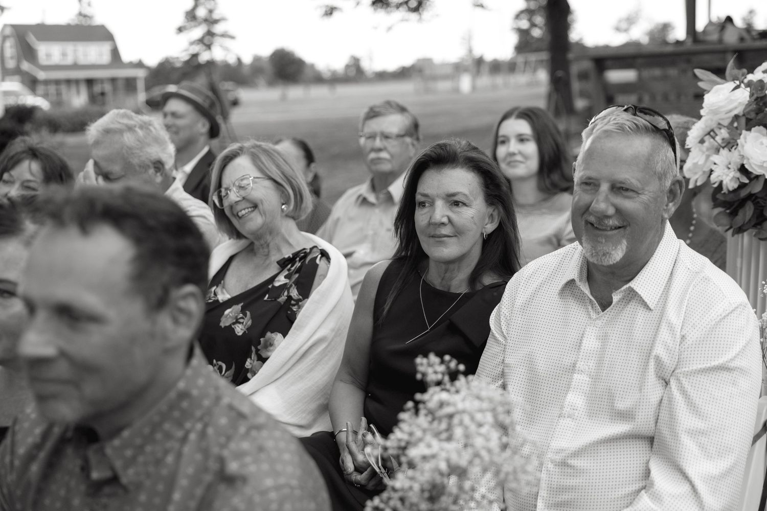 Black and white photo of smiling wedding guests seated outdoors during a countryside ceremony.