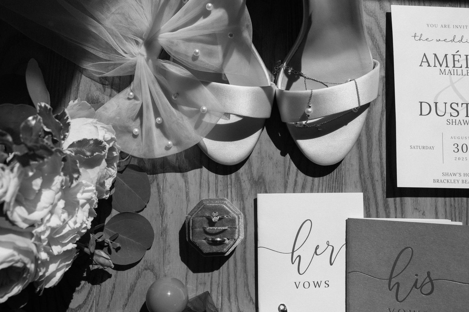 Black and white flat lay of wedding details including heels, vow books, rings, and invitation.