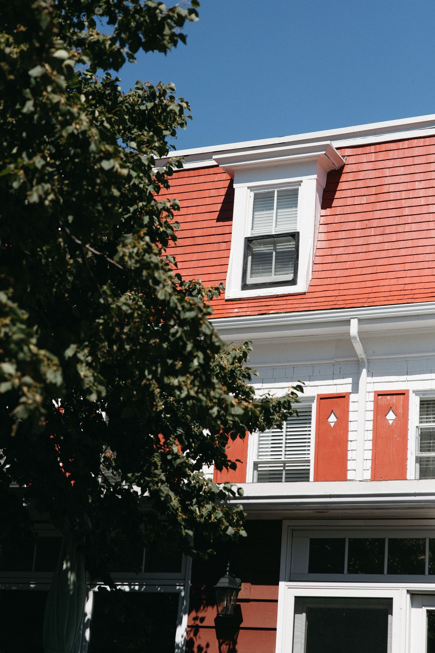 Red shingled colonial home with white trim and dormer window framed by green trees on a sunny day.