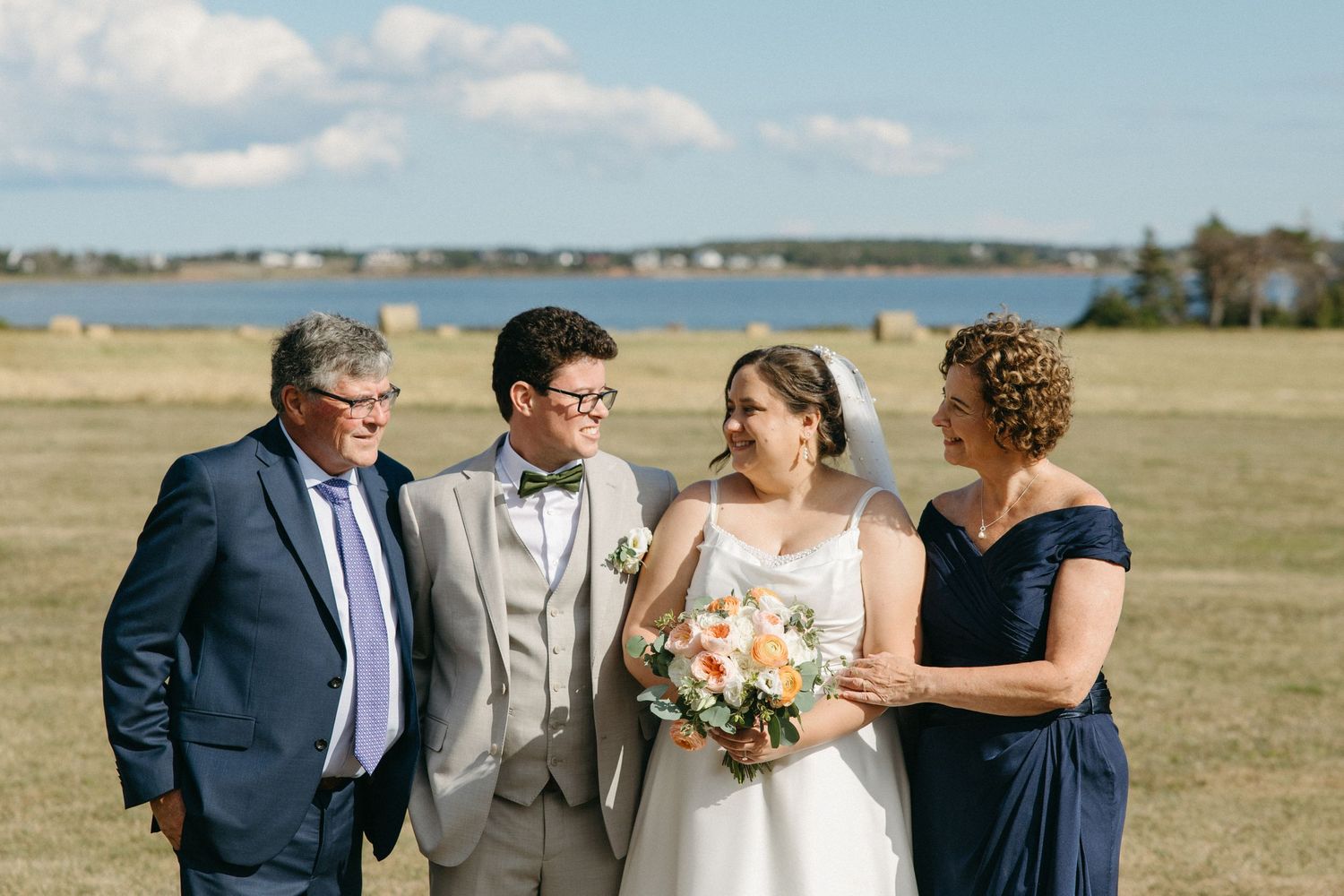 Bride and groom pose with family members in a sunny field with a scenic waterfront backdrop.