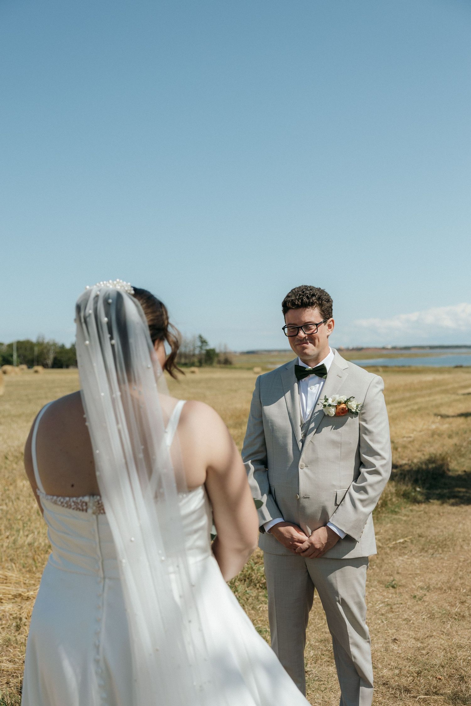 Groom in gray suit and bow tie smiling at veiled bride during romantic outdoor first look in sunny field.