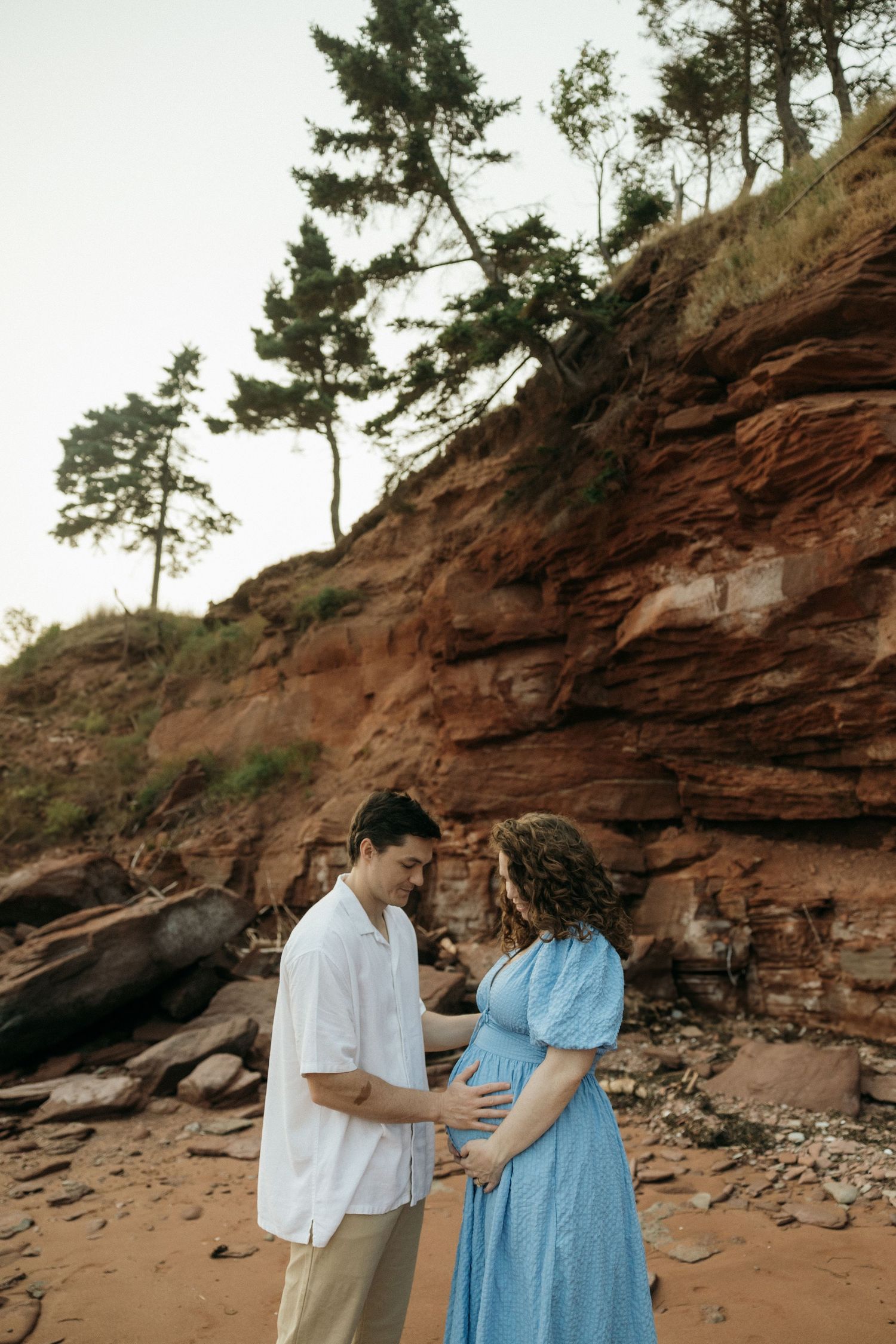 A romantic maternity photoshoot features a couple standing before red sandstone cliffs.