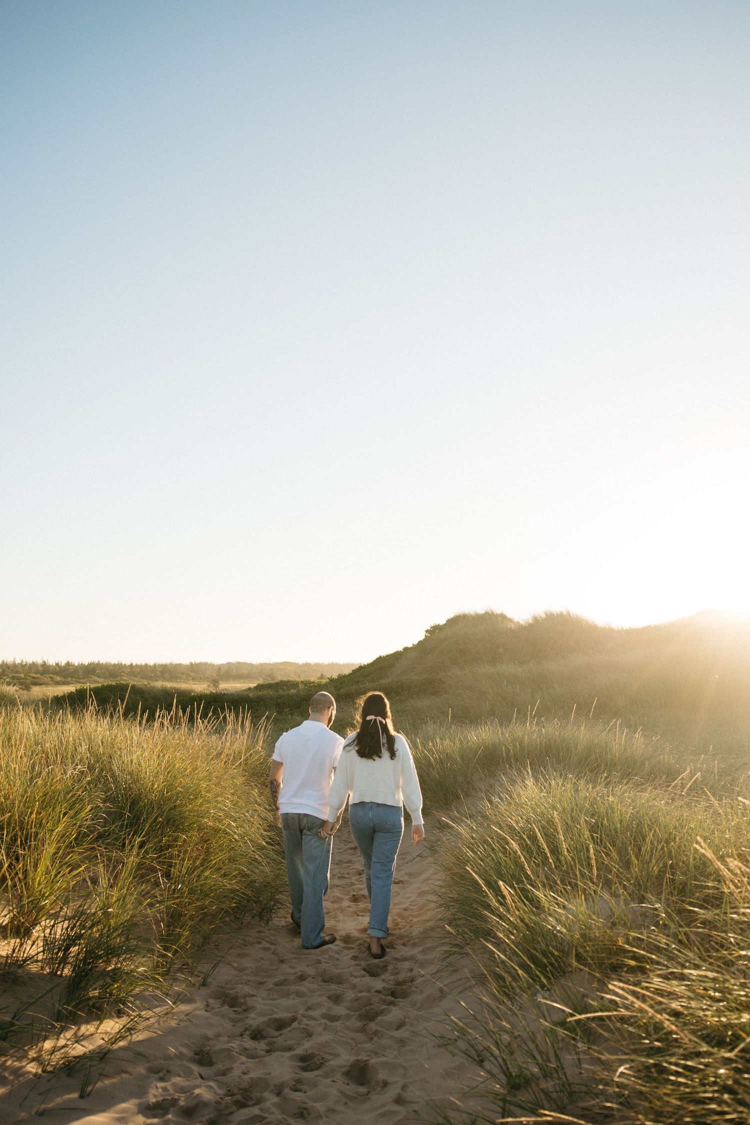 Couple walking hand in hand through beach grass at sunset.