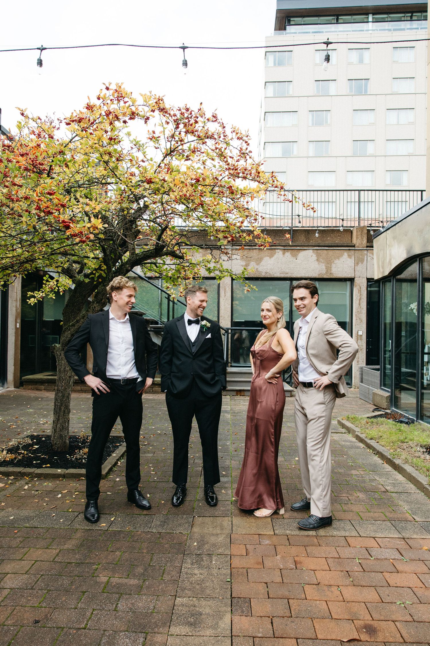 Four people in formal attire pose casually in a modern courtyard setting with autumn trees.