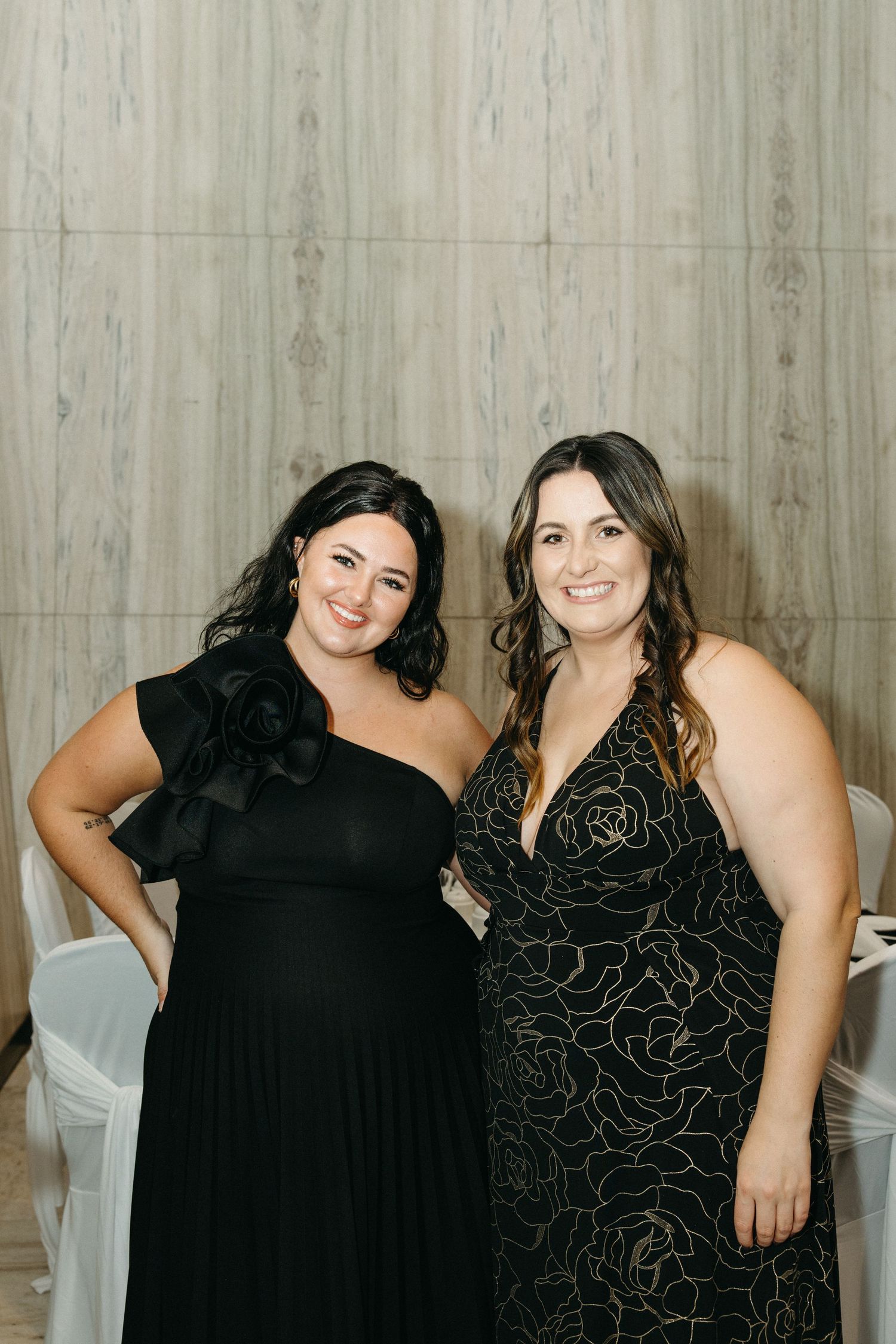 Two friends in elegant black dresses pose together against a light colored wall.