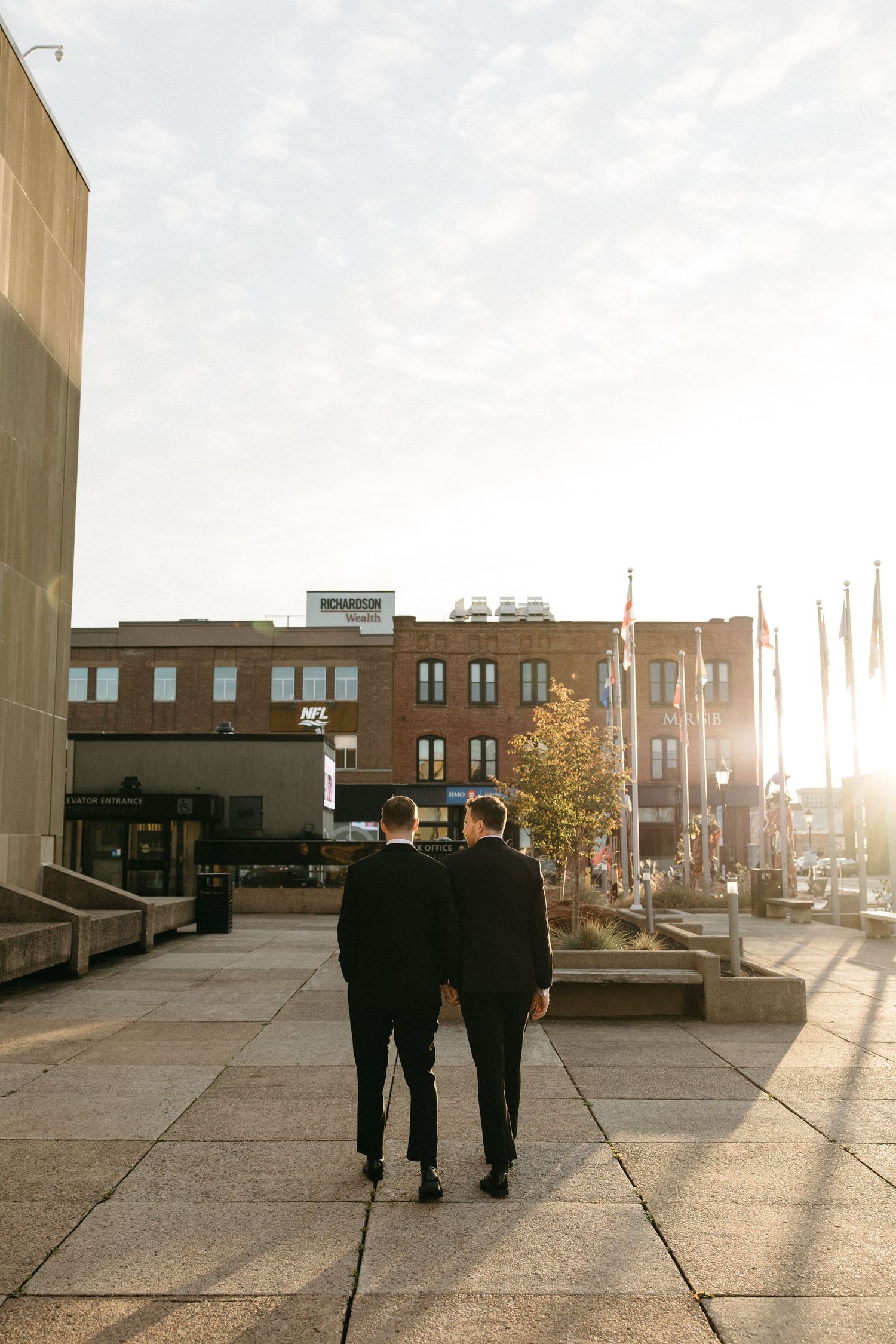 Silhouetted figures in formal attire walking away into golden sunset light on city sidewalk.