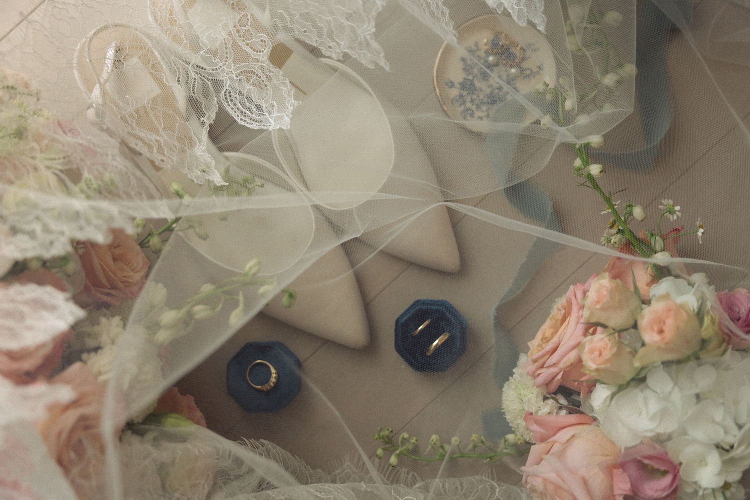 Wedding rings resting on a beige box surrounded by lace, pink roses, and bridal bouquet details.