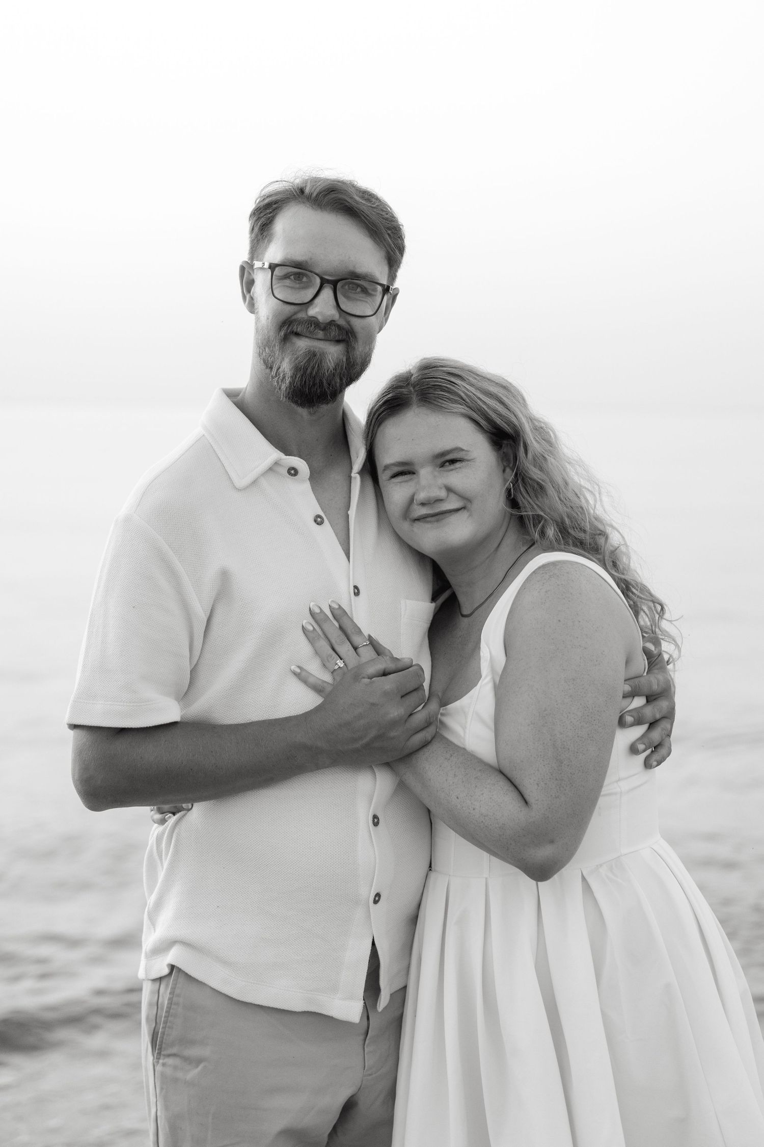 Black and white photo of a couple embracing on a beach, woman showing engagement ring, man wearing glasses.