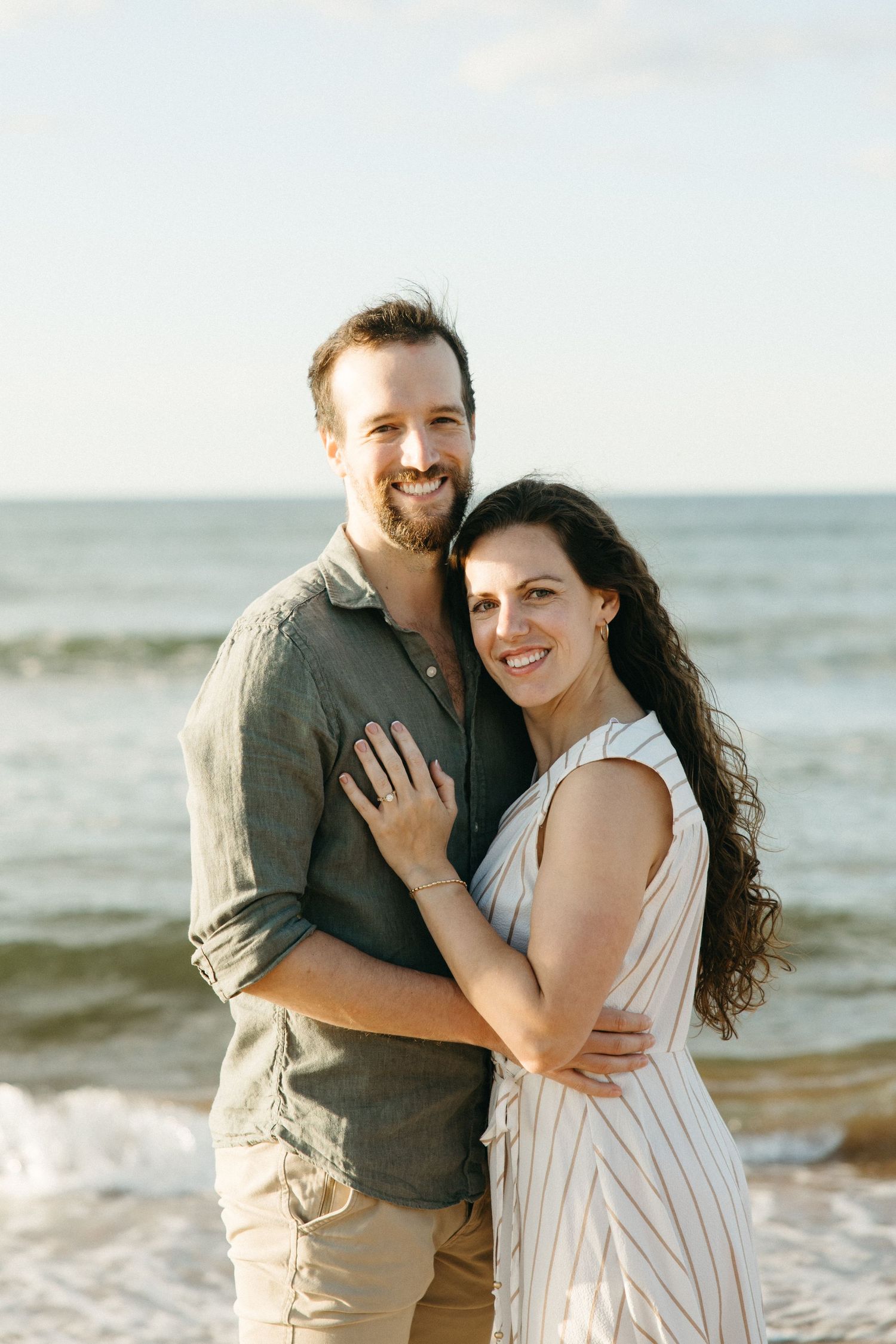 Happy couple embracing on beach with ocean waves behind them, woman in striped dress smiles warmly.