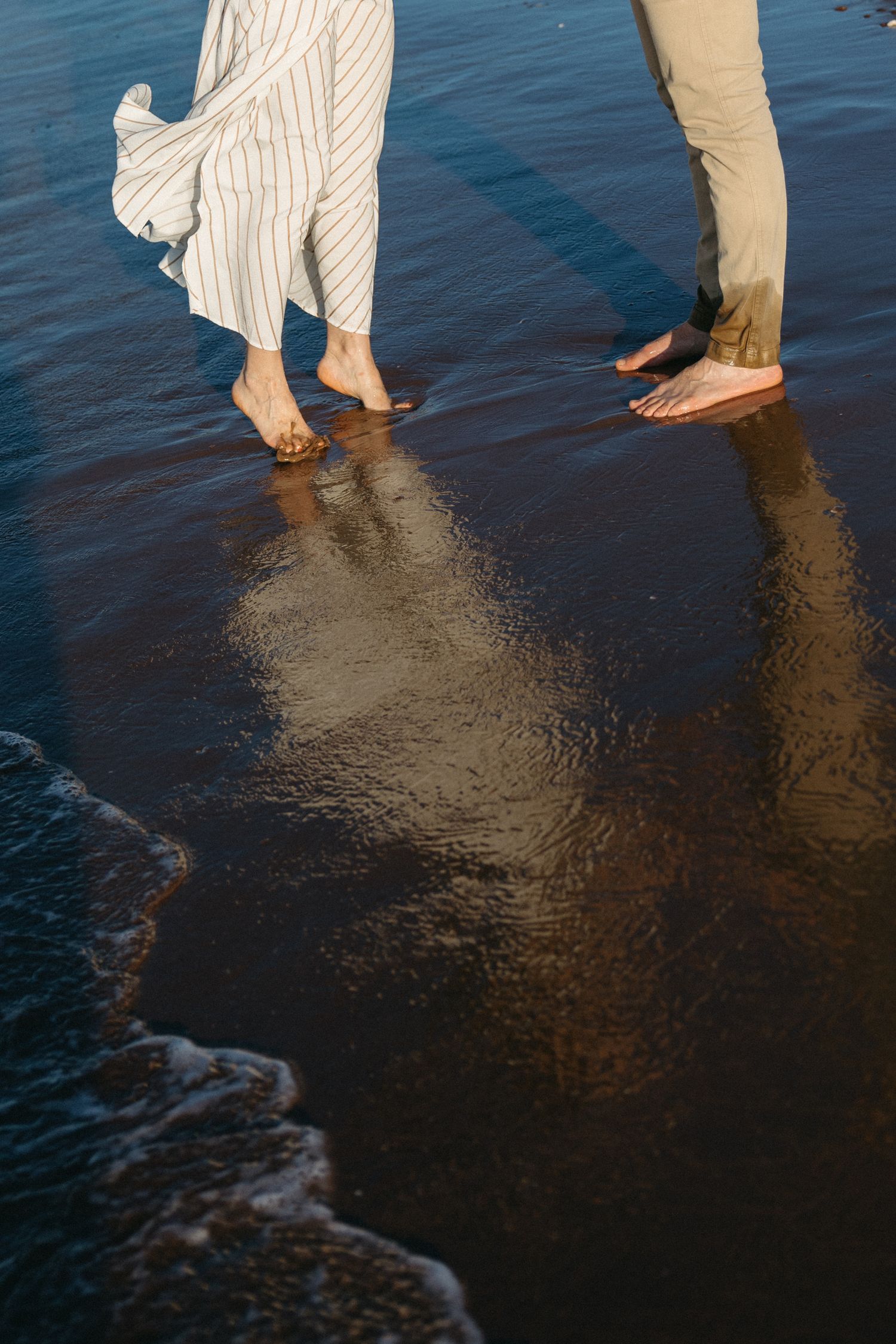 Barefoot couple walking on wet sandy beach shoreline with gentle waves and reflections.