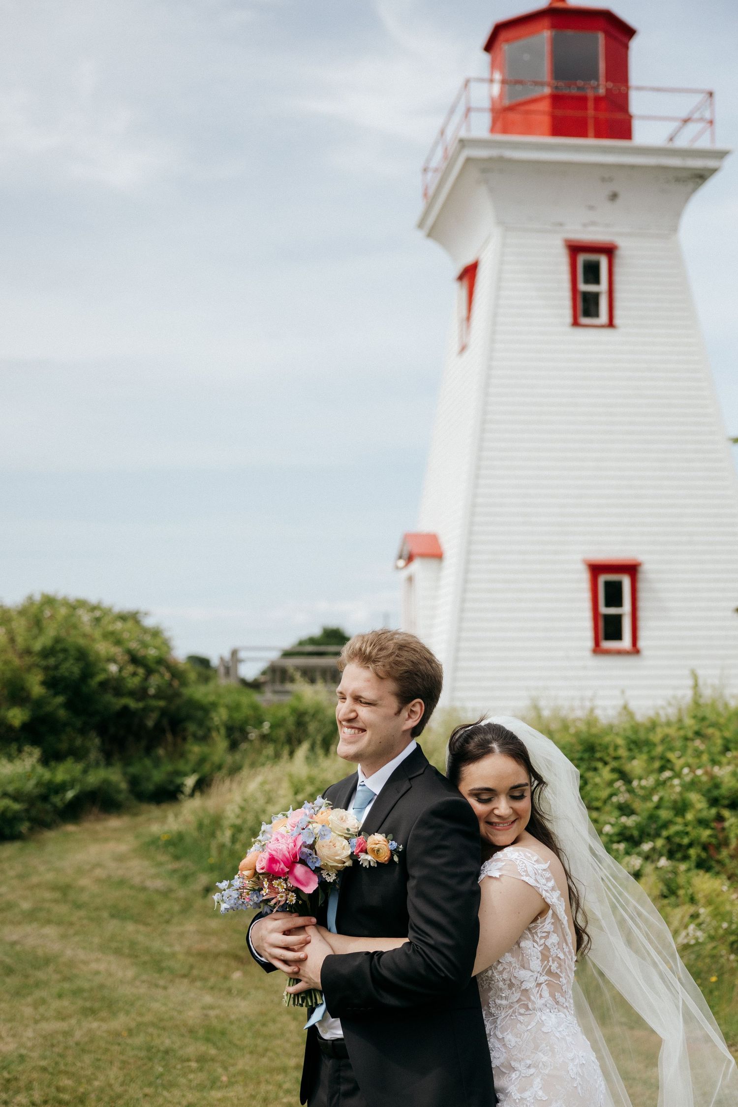 A romantic wedding portrait captured in front of a coastal lighthouse with red trim.