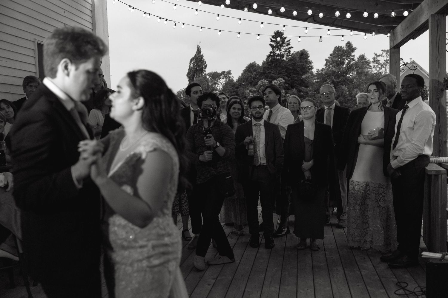 A couple dances on a deck while wedding guests look on during an evening reception.
