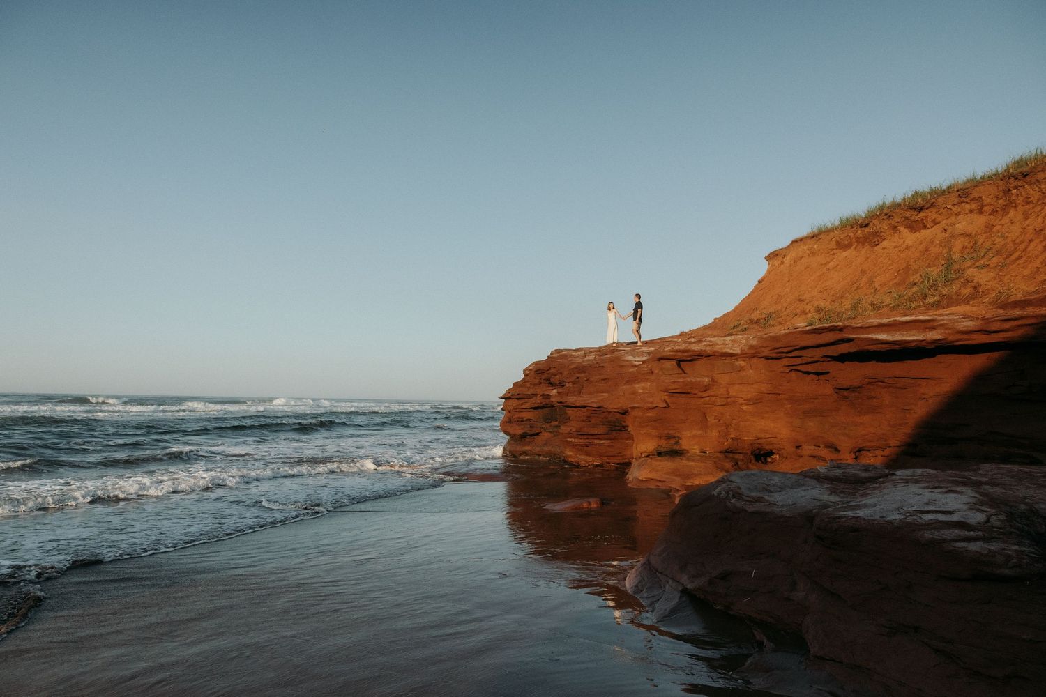 Two silhouettes stand atop red coastal cliffs overlooking the ocean at sunset.