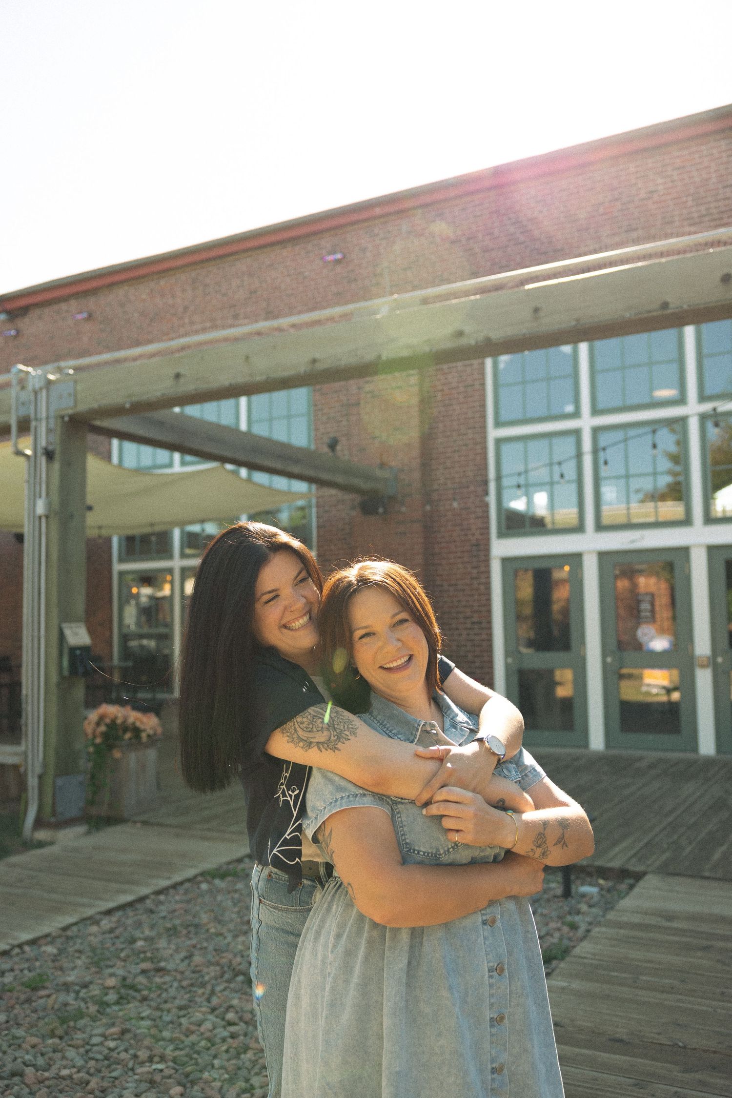 Two people hugging and laughing together on a sunny day outside a building.
