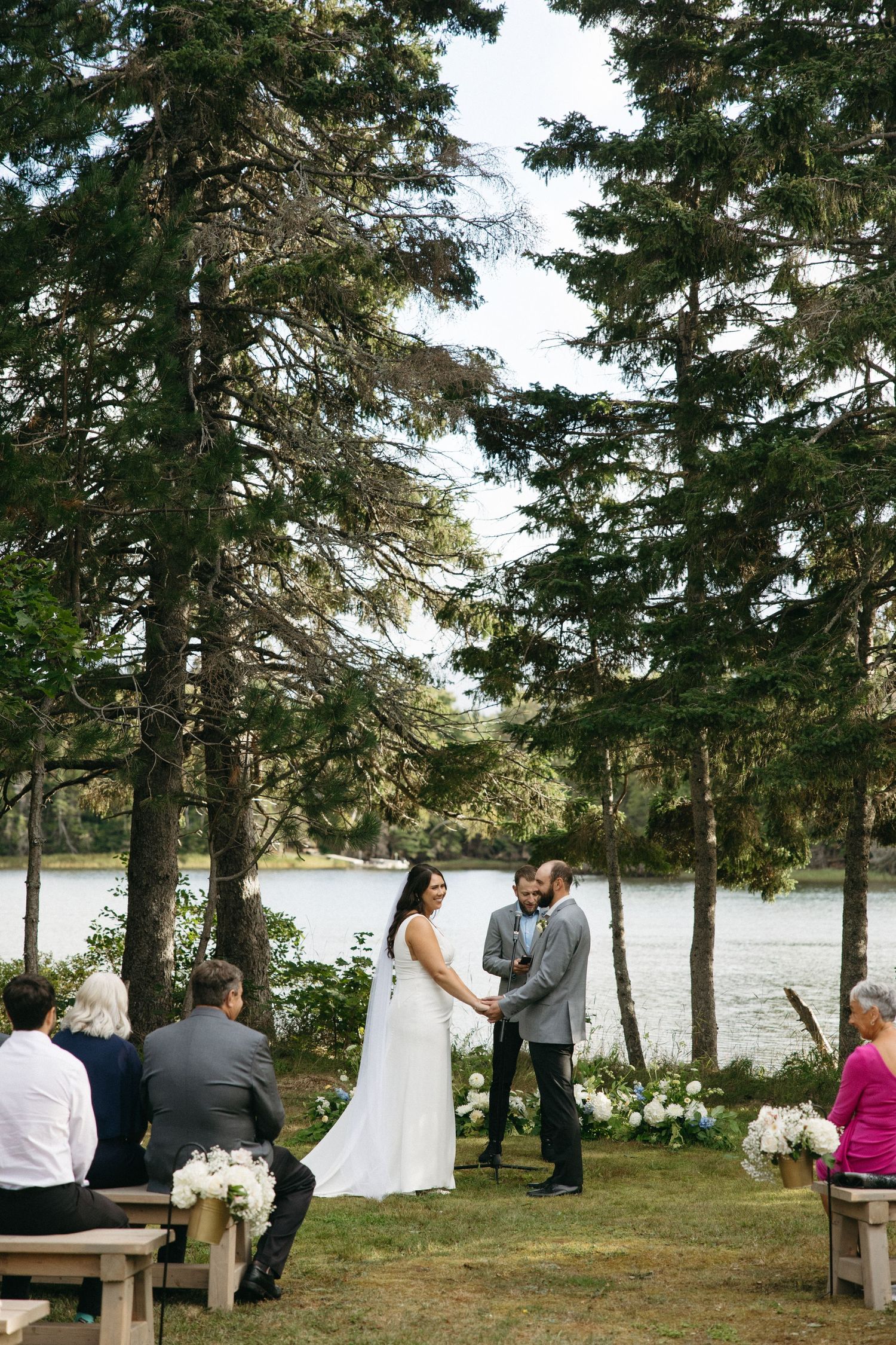 Wedding ceremony site with wooden benches arranged along a waterfront among tall pine trees.