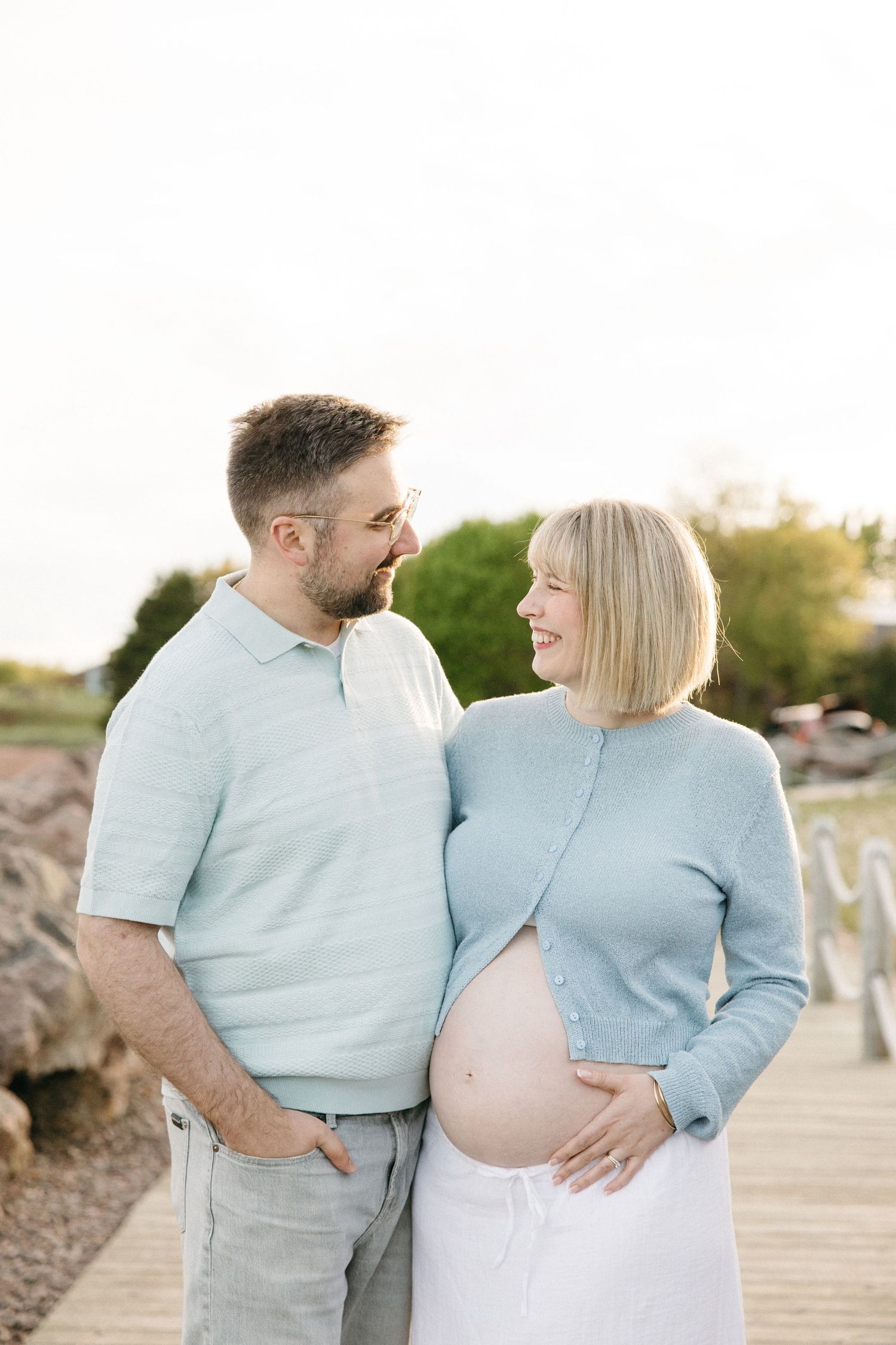 Happy expecting couple in matching blue outfits smile at each other on a sunny outdoor wooden boardwalk.