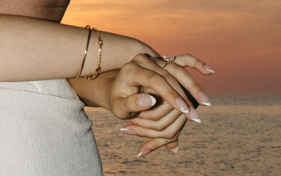 Close-up of clasped hands with gold bracelet and ring, elegant French manicure nails, sunset beach background.