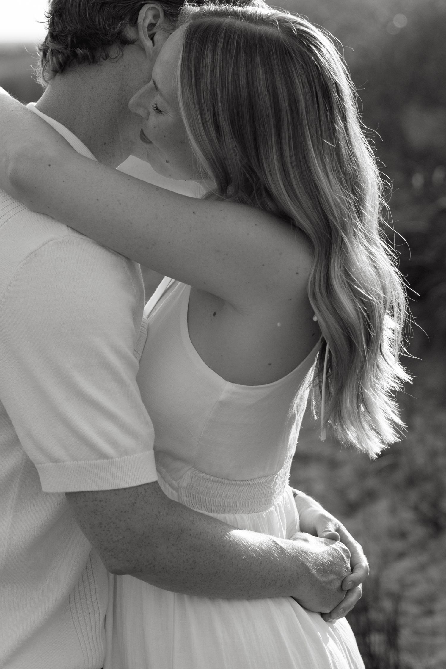 Black and white photo of couple embracing and kissing outdoors, woman in white dress with long blonde hair.