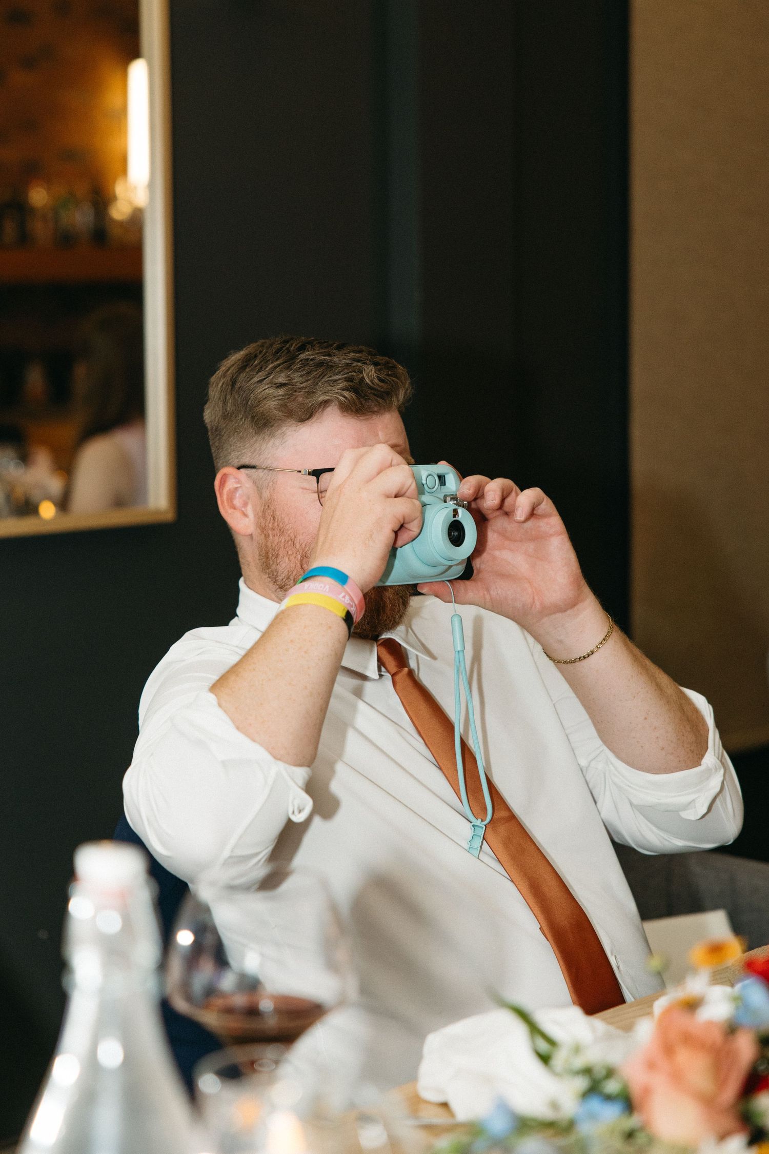 Man in a white shirt and rust-colored tie takes a photo with a turquoise instant camera at a reception table with flowers and glassware.