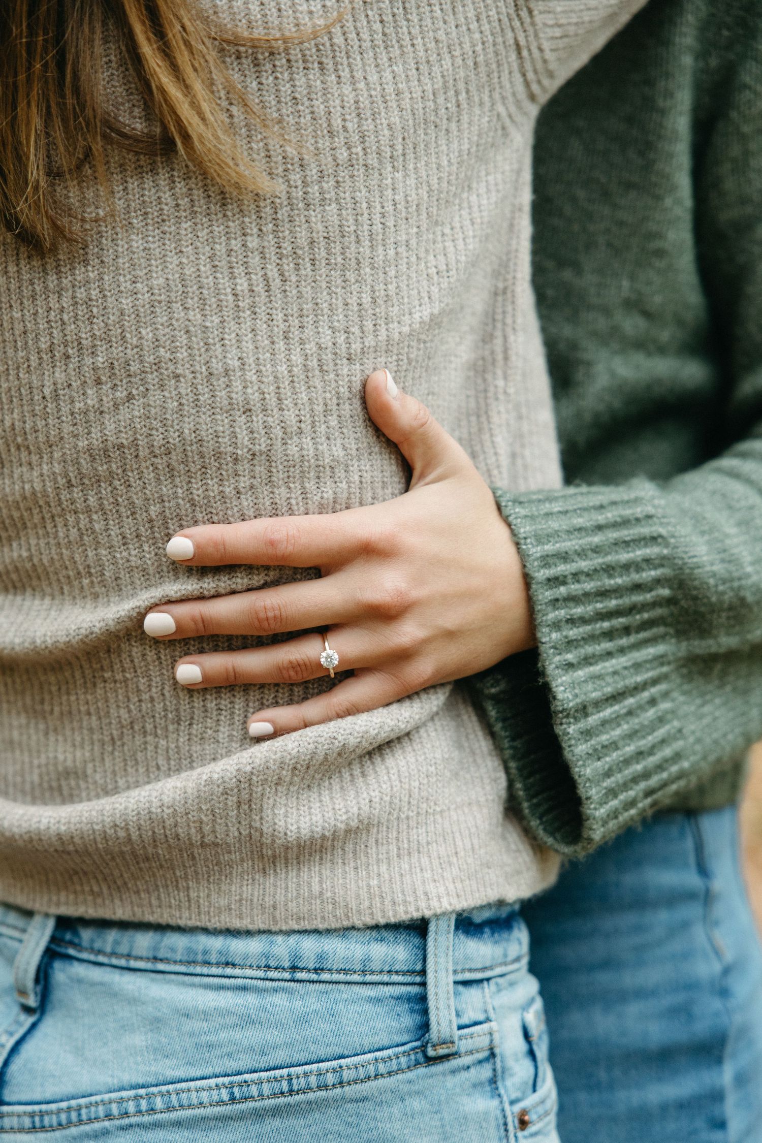 Close-up of a woman's hand with a diamond engagement ring resting on a partner's waist, both wearing cozy knit sweaters and jeans.