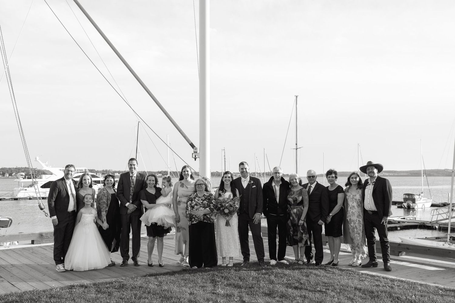 Black and white photo of a wedding party standing together on a marina dock with sailboats and masts in the background.