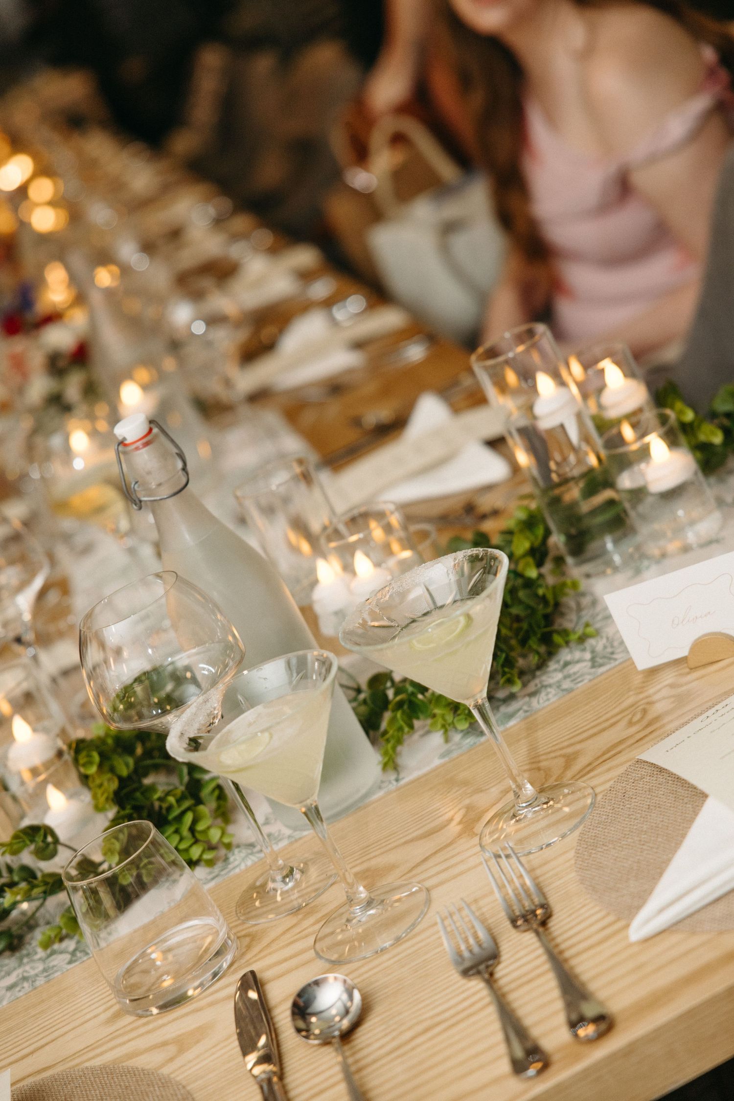 Elegant wedding reception table with candlelight, greenery garnish, cocktail glasses, vintage flatware and a guest in a pink dress.