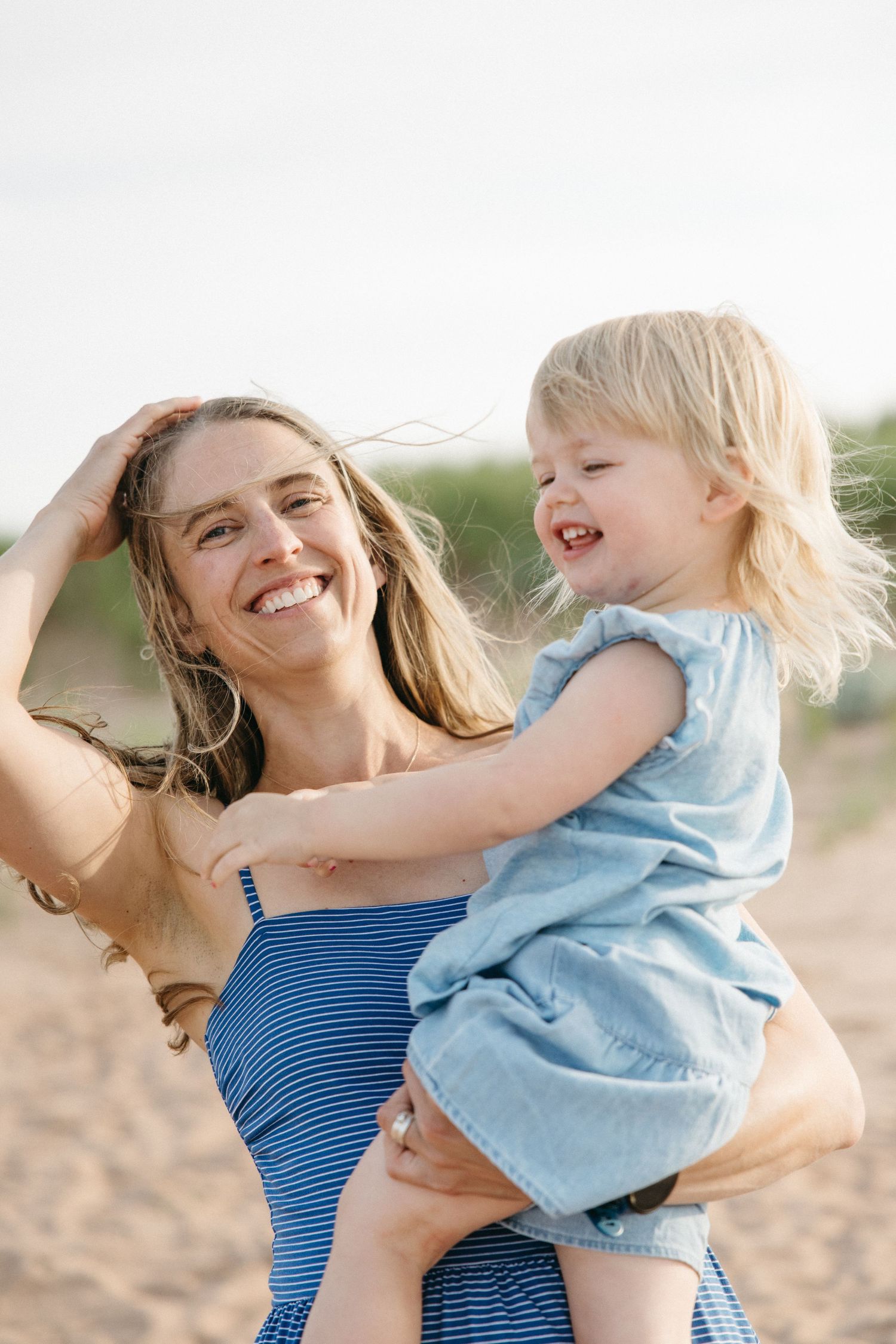 Smiling mother holding laughing blonde toddler girl on a sandy beach, both wearing blue outfits.