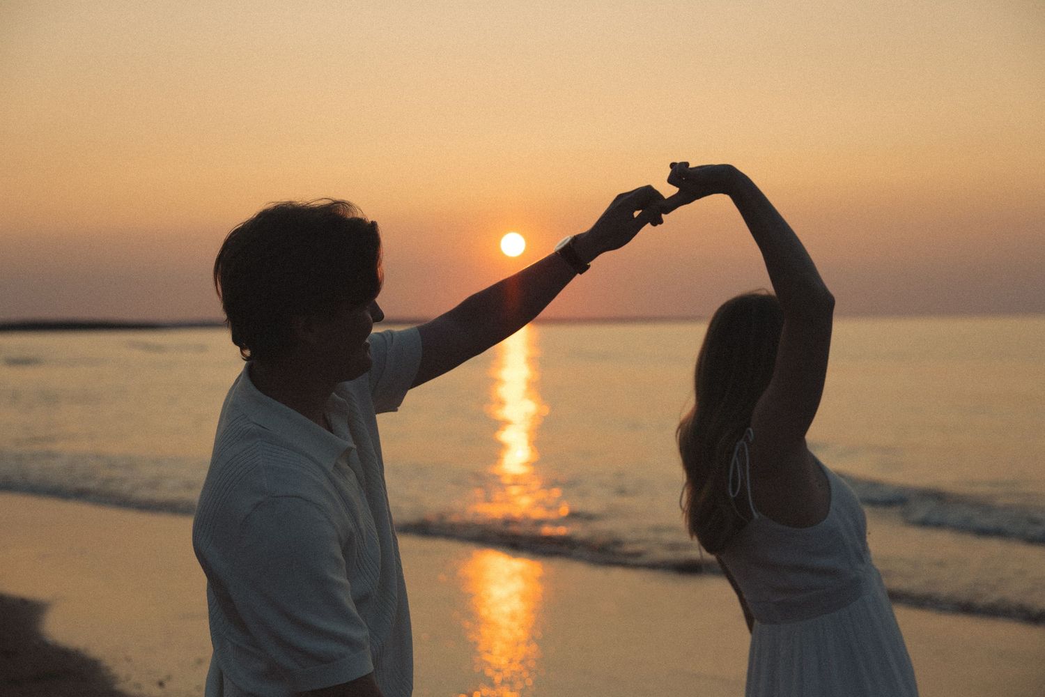 Couple dancing on beach at sunset, man spinning woman under golden sun over ocean horizon.