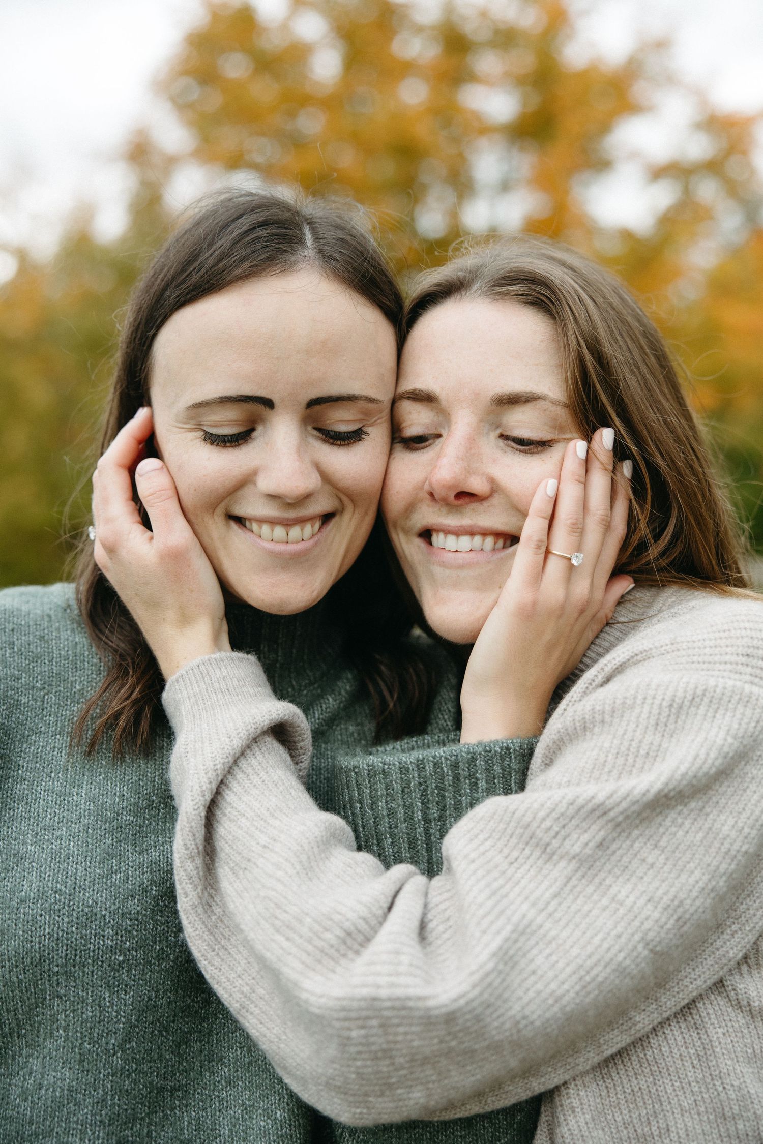 Two smiling women with closed eyes embrace cheek to cheek in cozy sweaters against a blurred autumn backdrop of golden fall foliage.