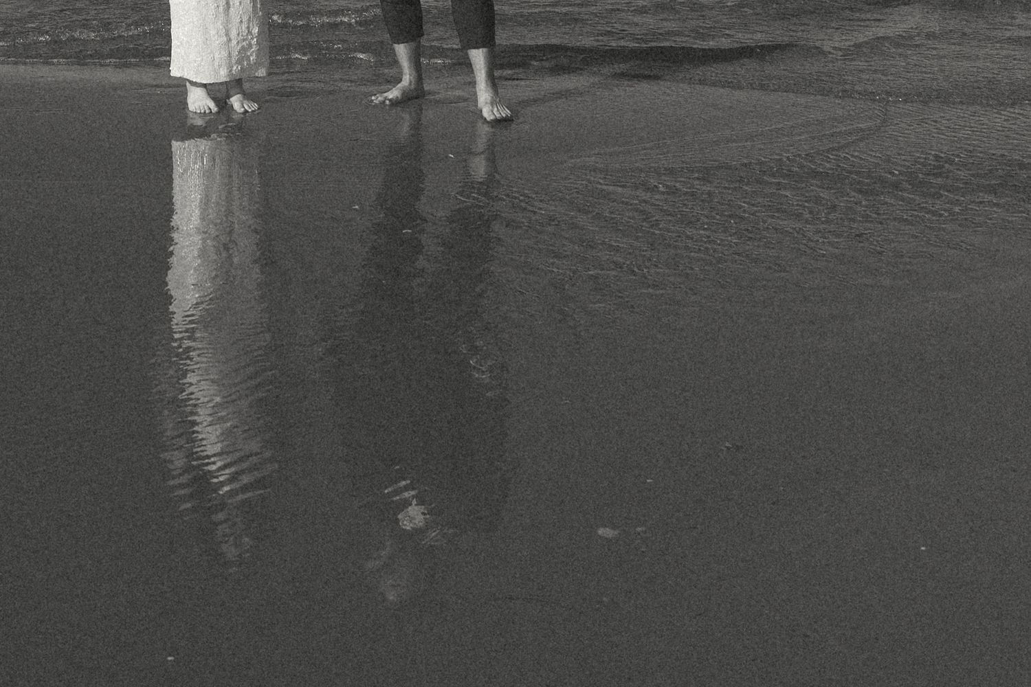 Black and white photo of two people standing barefoot in shallow water at the beach, their legs and reflections mirrored on the wet sand.