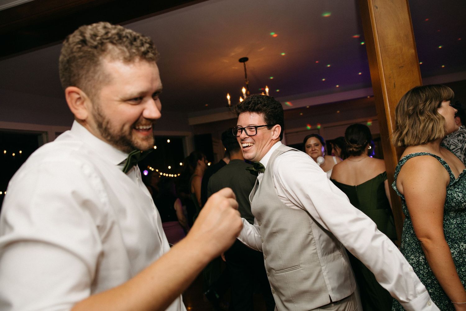 Two men in suits laughing and dancing together at wedding reception.