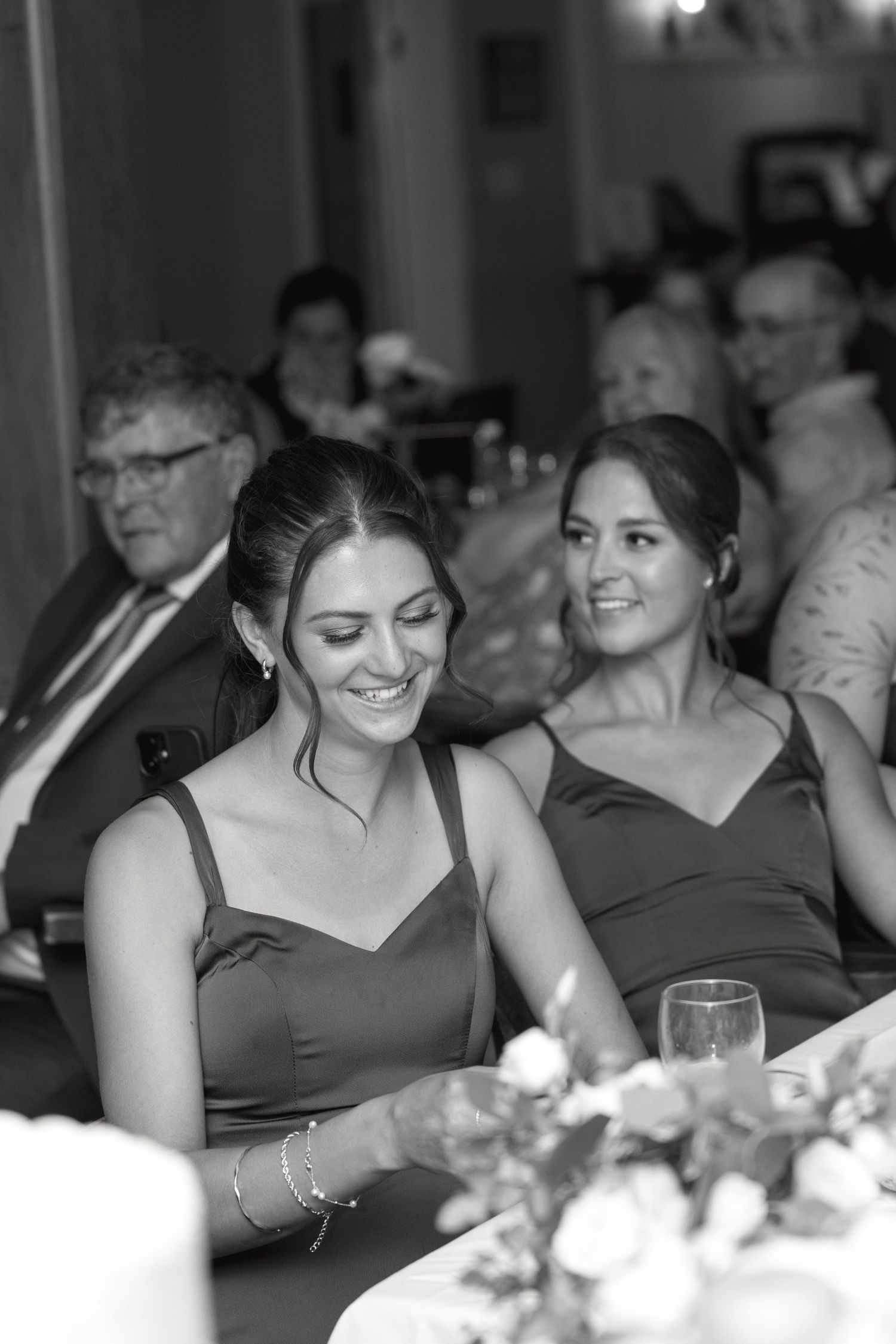Two bridesmaids in dark green dresses smile and laugh during wedding reception speeches in black and white.