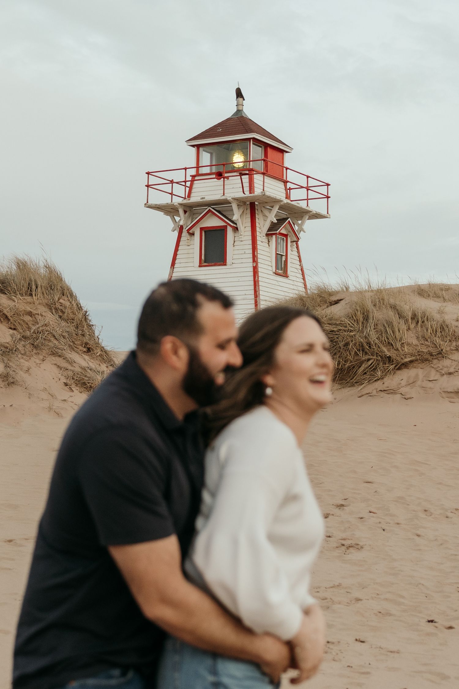 Couple embracing near a red and white lighthouse on a sandy beach under cloudy skies.