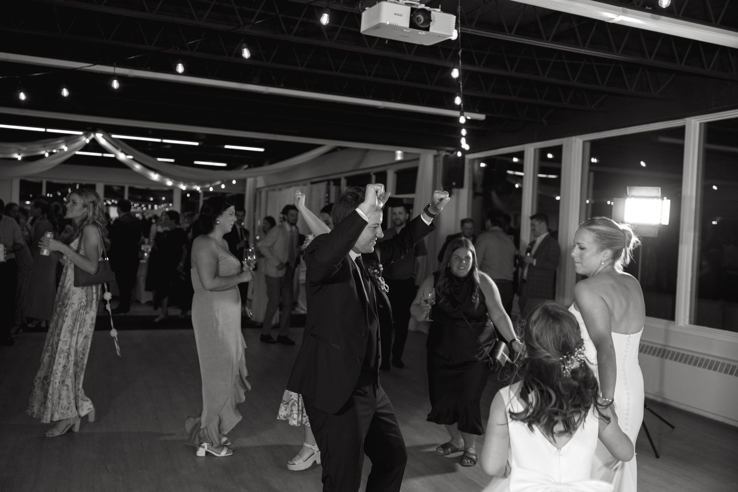Guests dance and celebrate at a wedding reception in a modern venue with exposed ceiling and string lights.
