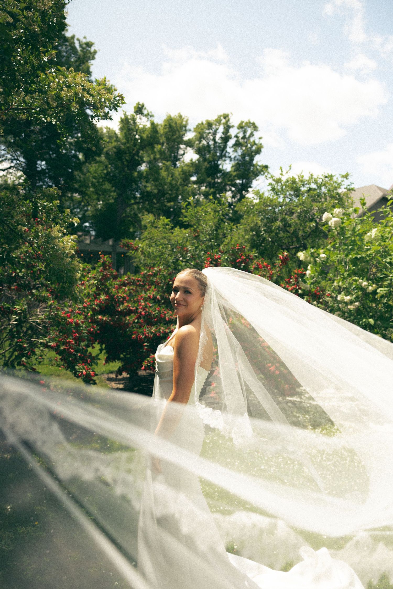 A long white wedding veil flows dramatically in the wind against a lush garden landscape.