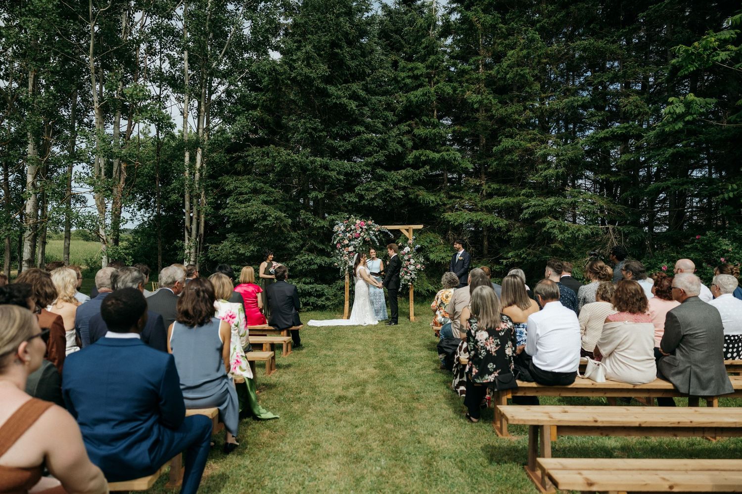 Guests seated on wooden benches during outdoor forest wedding ceremony.