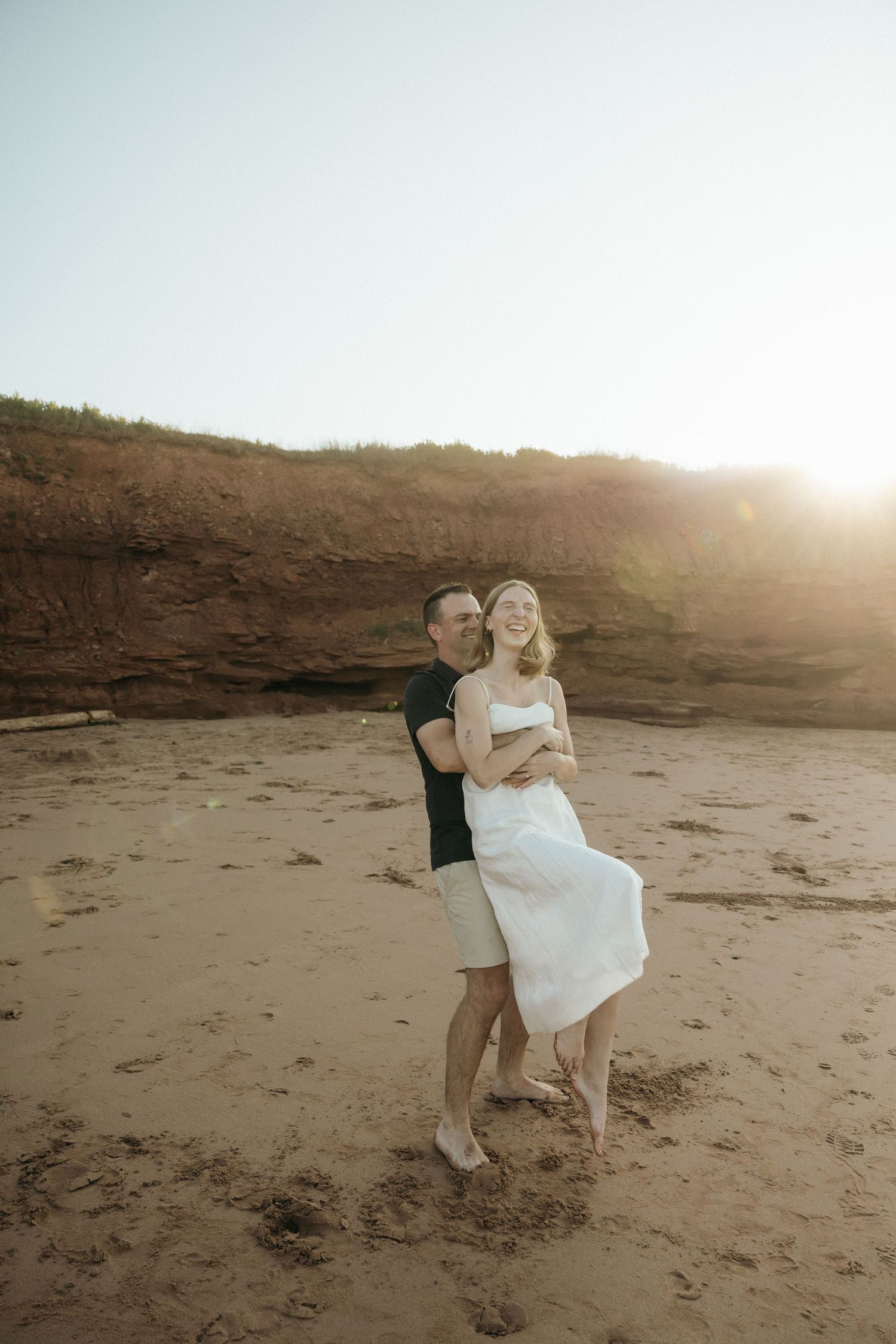 Couple embracing on sandy beach with cliffs and sunset in background.