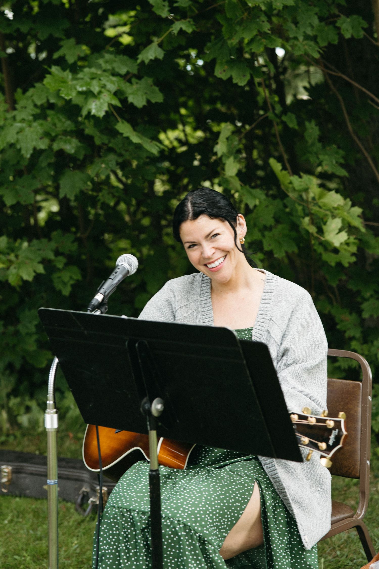 Musician performs at outdoor event with music stand and microphone under green foliage.