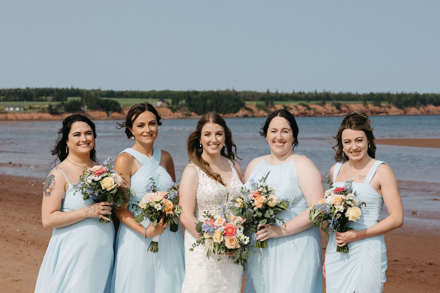 Bridesmaids in light blue dresses holding floral bouquets on beach.